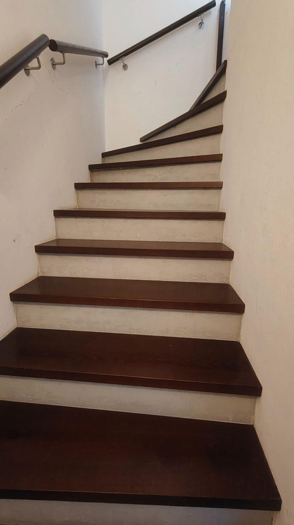 Klatka schodowa z drewnianymi stopniami i pomalowanymi na biało ścianami. Widoczne ślady po szpachlowaniu na ścianach. Ujęcie z dołu, podkreślające pionowy układ schodów.