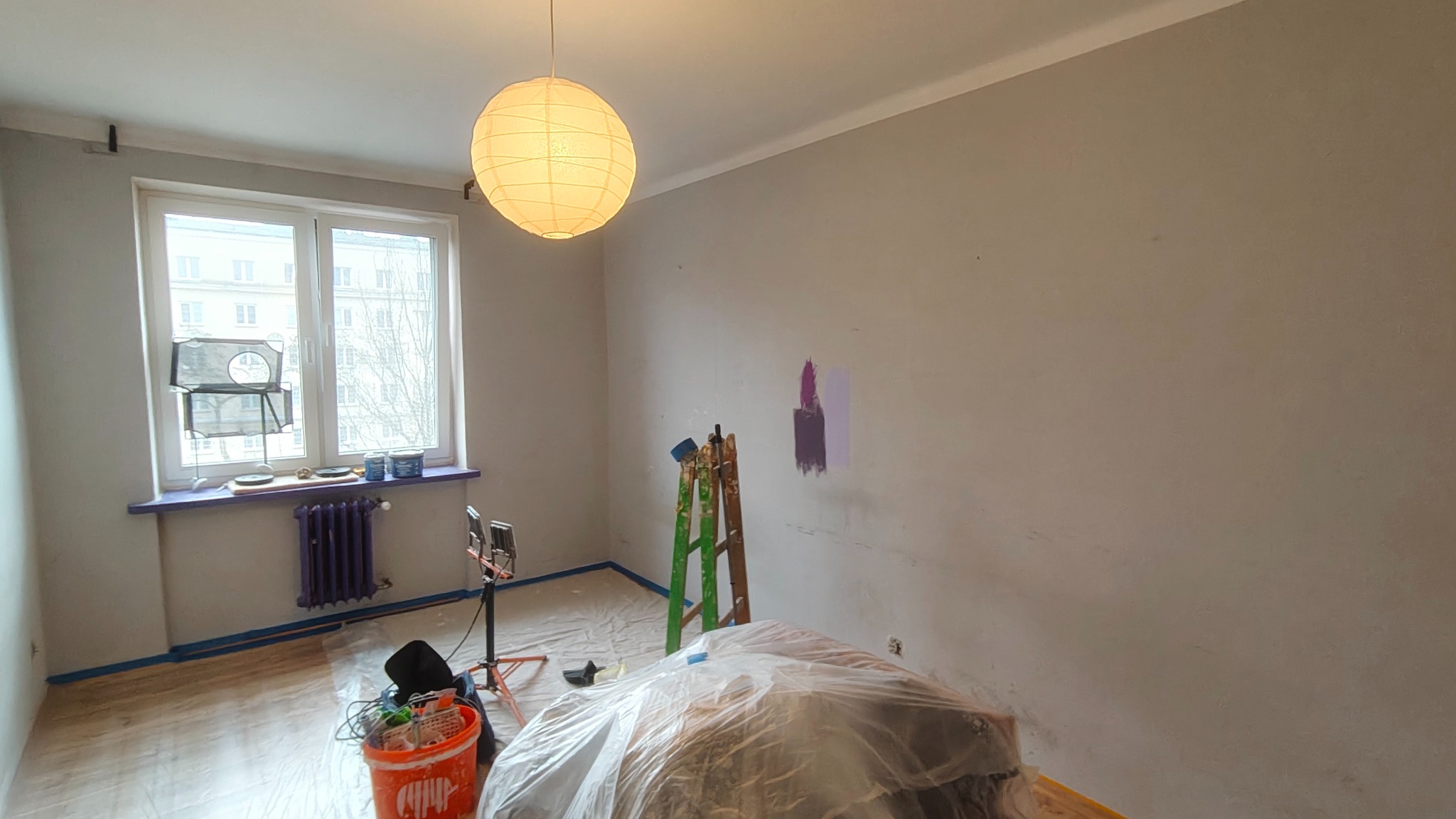 Pokój w trakcie malowania: zabezpieczone podłogi, drabina, lampa wisząca, ślady testowych kolorów na ścianie. Widoczne narzędzia i farby. Przygotowania do odświeżenia wnętrza.