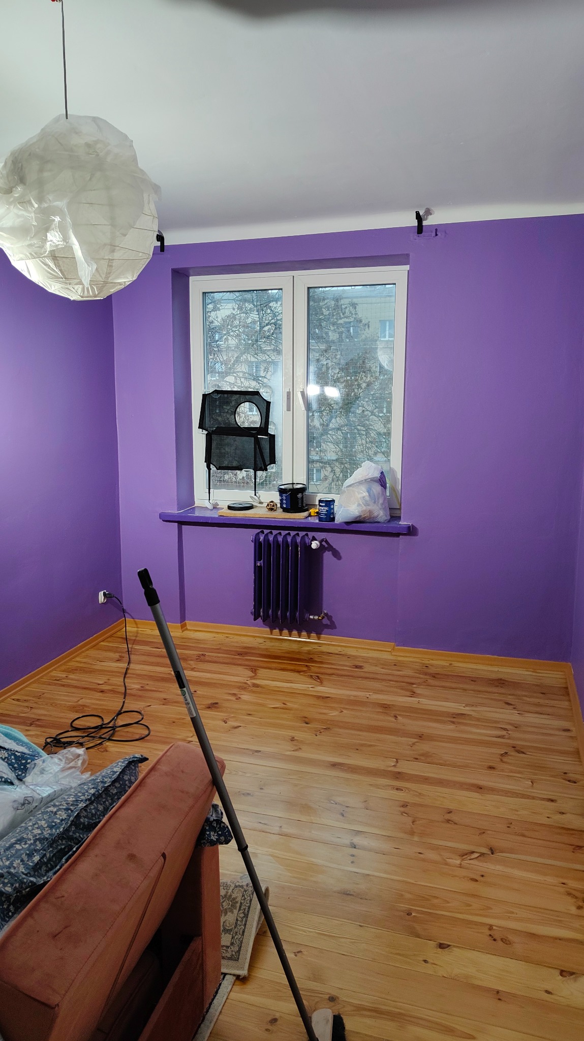 Pokój po malowaniu ścian na fioletowo. Widoczne okno, grzejnik, lampa i narzędzia malarskie. Podłoga drewniana, częściowo zakryta meblami i folią.