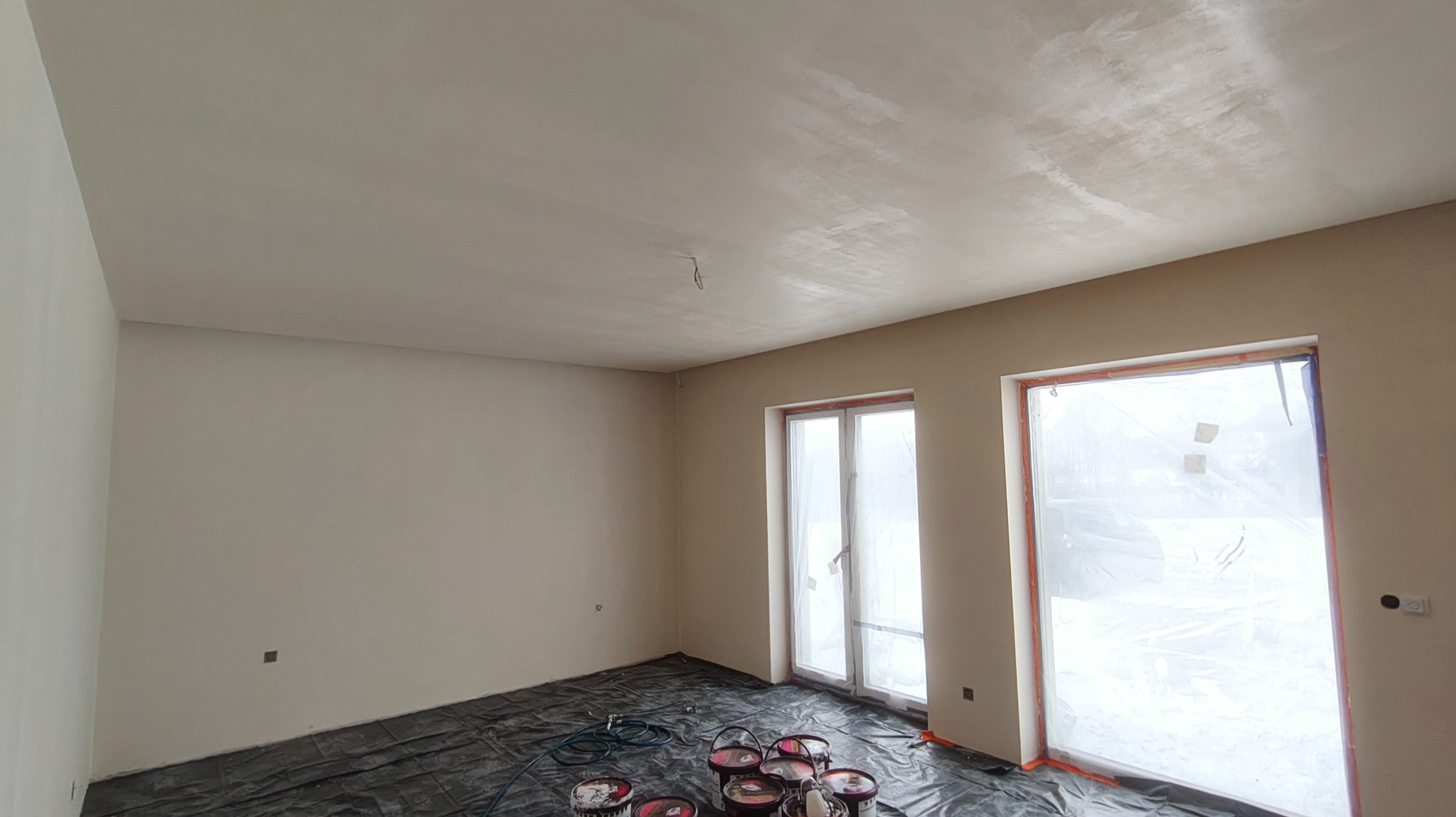 Wnętrze po szpachlowaniu i gruntowaniu ścian, przygotowane do malowania. Podłoga zabezpieczona folią, widoczne puszki z farbą. Ściany w jasnym odcieniu beżu.