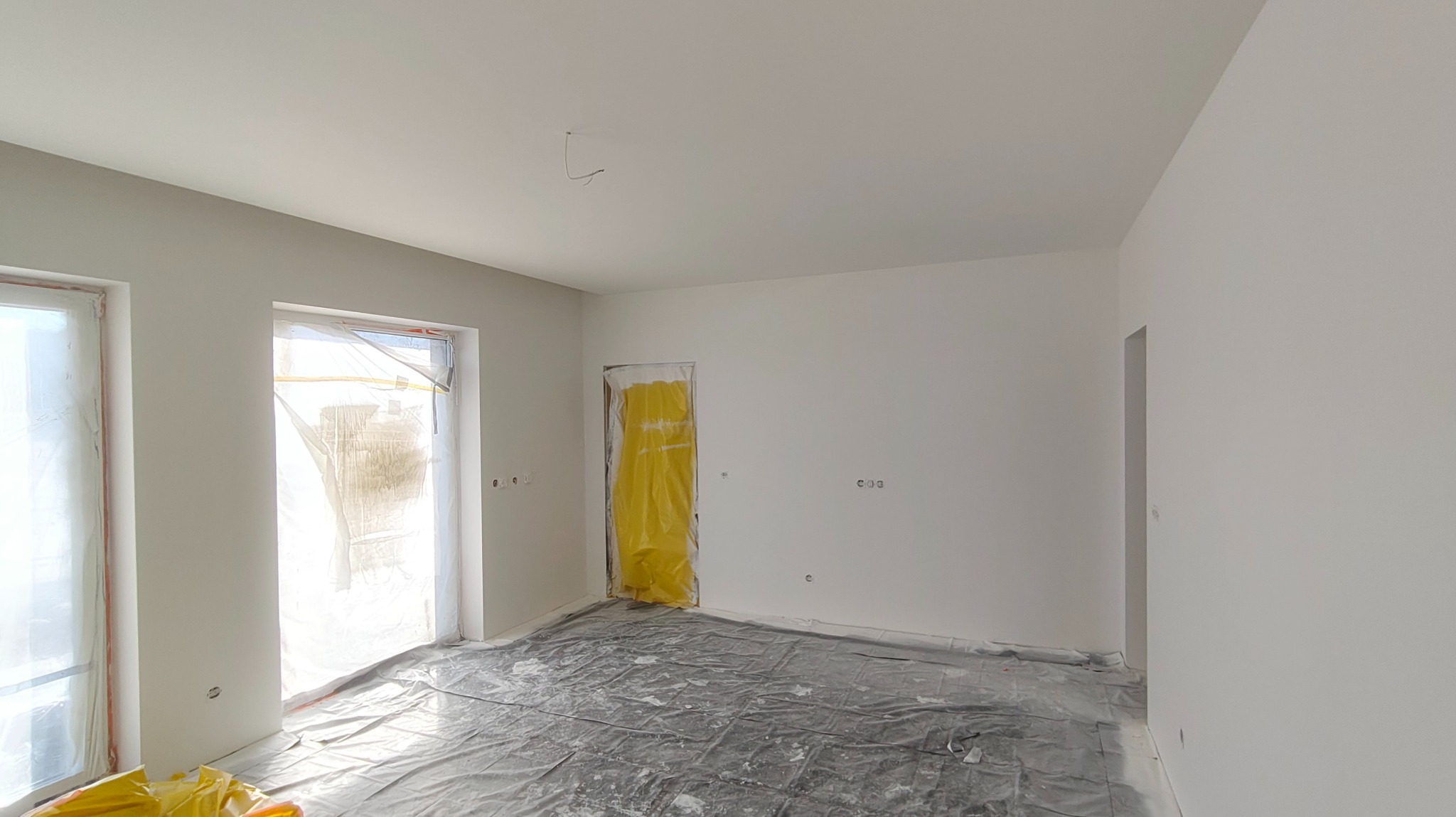 Białe ściany po malowaniu natryskowym w pustym pokoju, zabezpieczone okna i drzwi folią malarską, podłoga przykryta folią ochronną. Widoczne ślady farby.