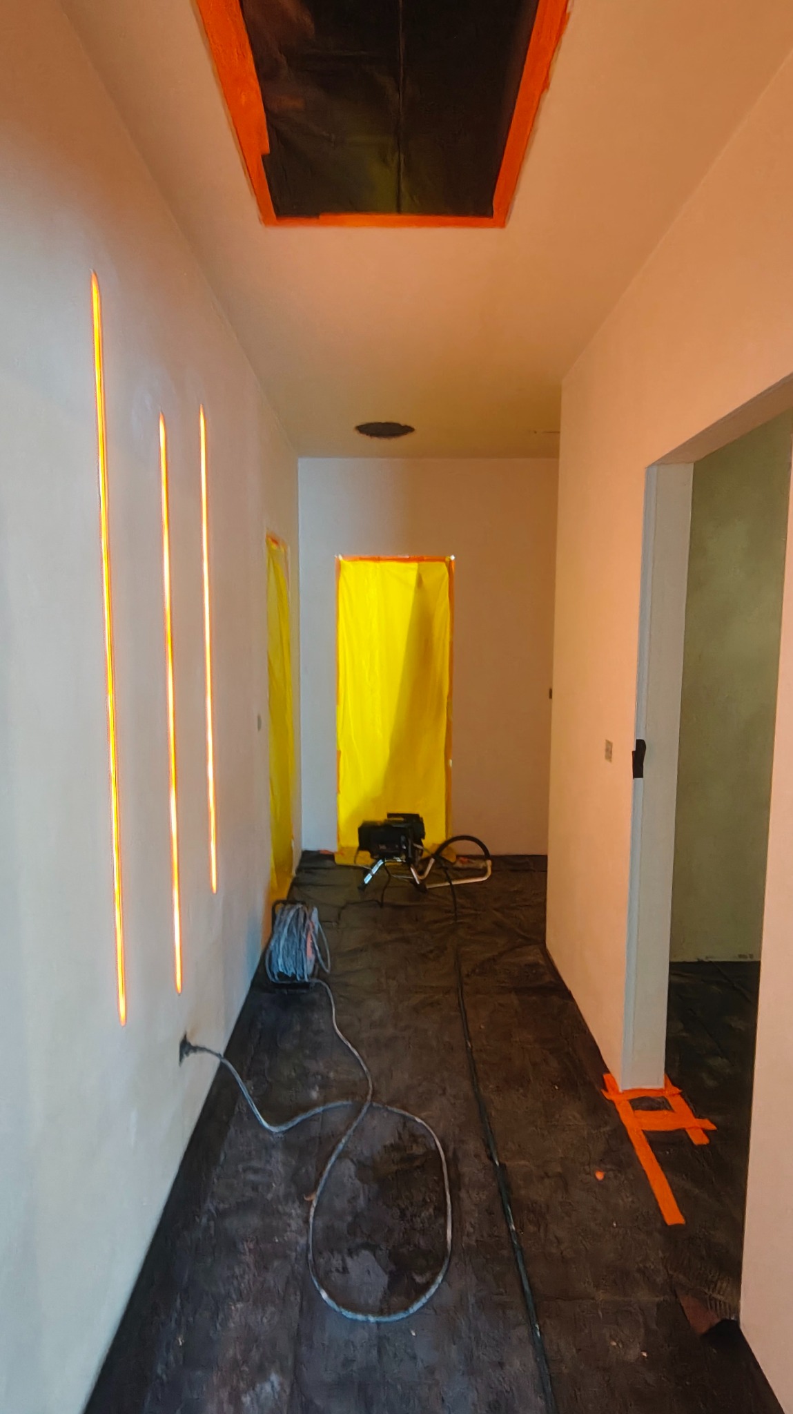 Przygotowane do prac wykończeniowych wnętrze z zabezpieczonymi otworami i podłogą. Widoczne szpary oświetleniowe w ścianie i sprzęt budowlany.