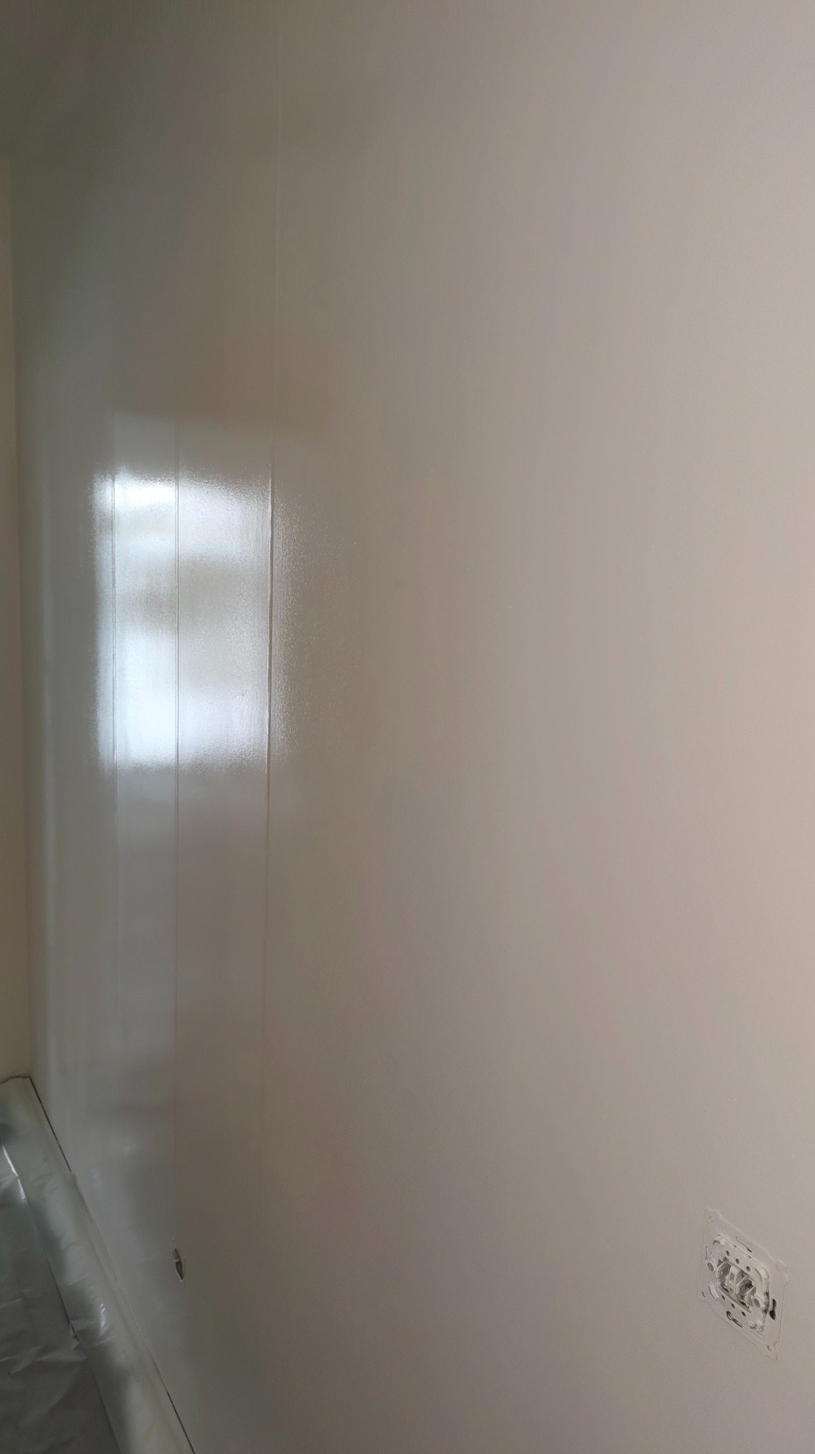 Świeżo pomalowana, gładka ściana w odcieniach bieli z widocznym gniazdkiem elektrycznym i folią ochronną na podłodze. Widoczne refleksy światła na powierzchni.