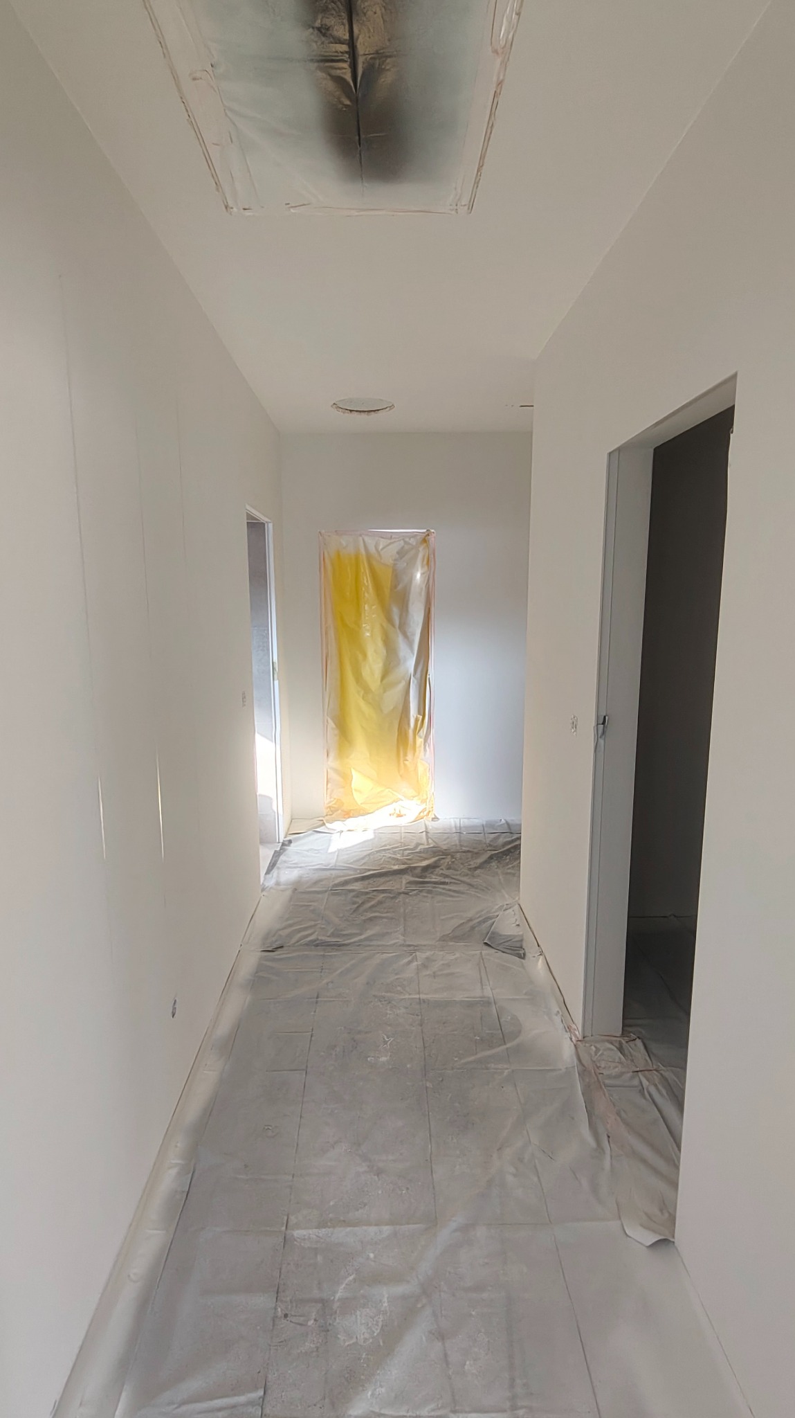 Korytarz w trakcie malowania natryskowego: białe ściany, zabezpieczona folią podłoga i drzwi. Widoczne otwory w suficie, jeden zabezpieczony folią.