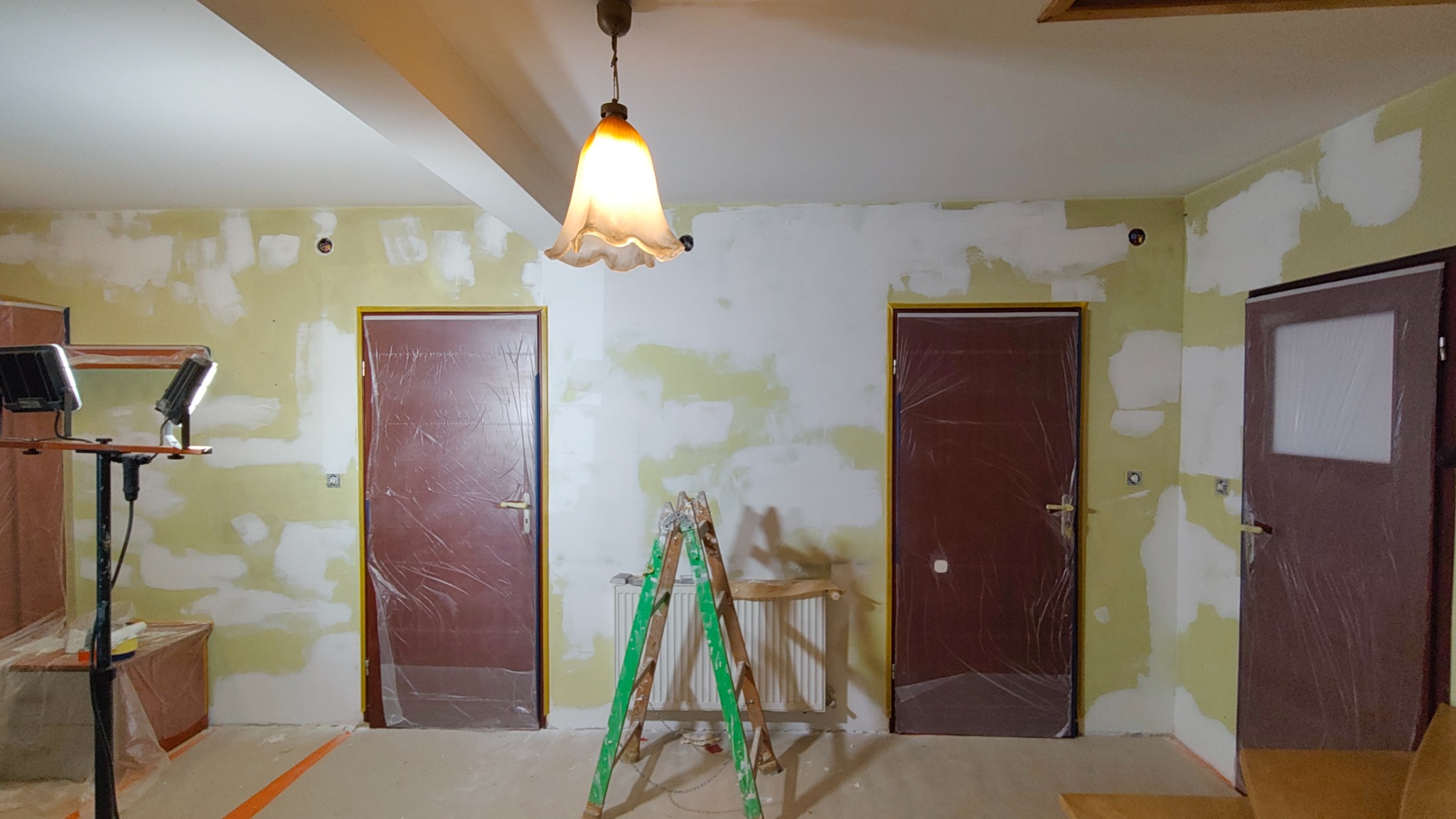 Pokój w trakcie prac wykończeniowych: ściany z nałożoną gładzią, zabezpieczone folią drzwi, drabina oparta o grzejnik, lampa wisząca. Widoczne ślady po szpachlowaniu.