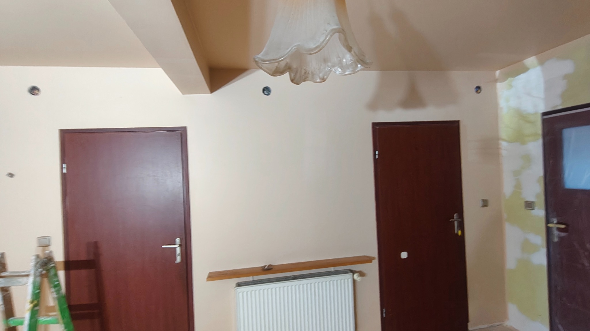 Pokój po szpachlowaniu i malowaniu, widoczne ślady po łataniu ścian, przygotowane do dalszych prac wykończeniowych. Dwie brązowe drzwi i biały grzejnik.
