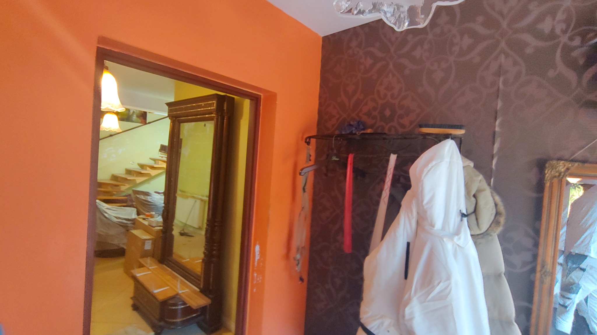 Pomarańczowa ściana z widocznym wejściem do pokoju w trakcie remontu. Widoczne zabezpieczenia i narzędzia. Na ścianie wieszak z ubraniami i lustro.
