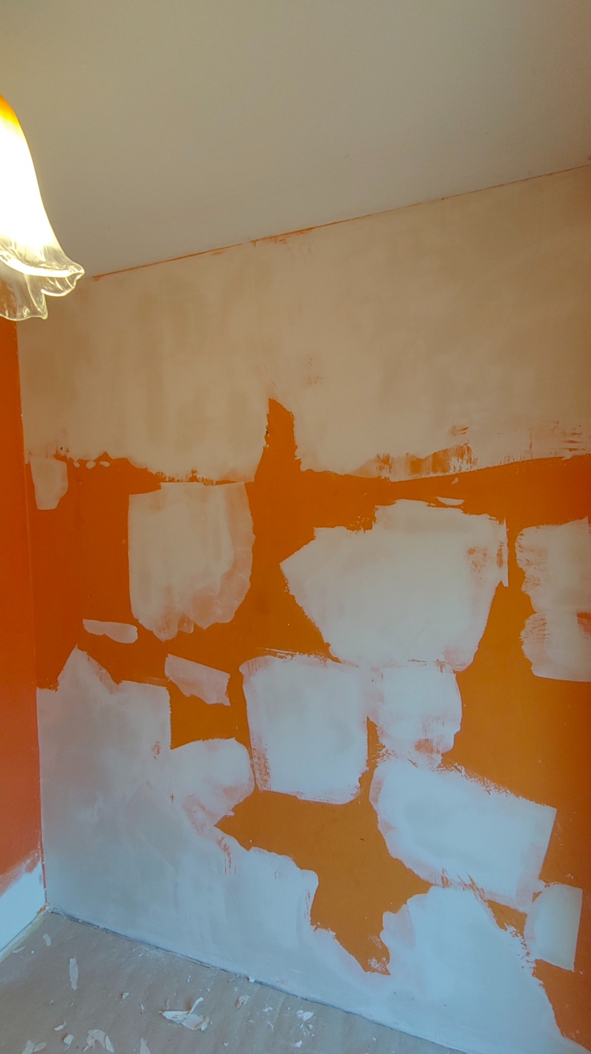Pomarańczowa ściana w trakcie szpachlowania, widoczne białe plamy gładzi. U dołu fragment podłogi, u góry lampa. Ściana przygotowywana do malowania.