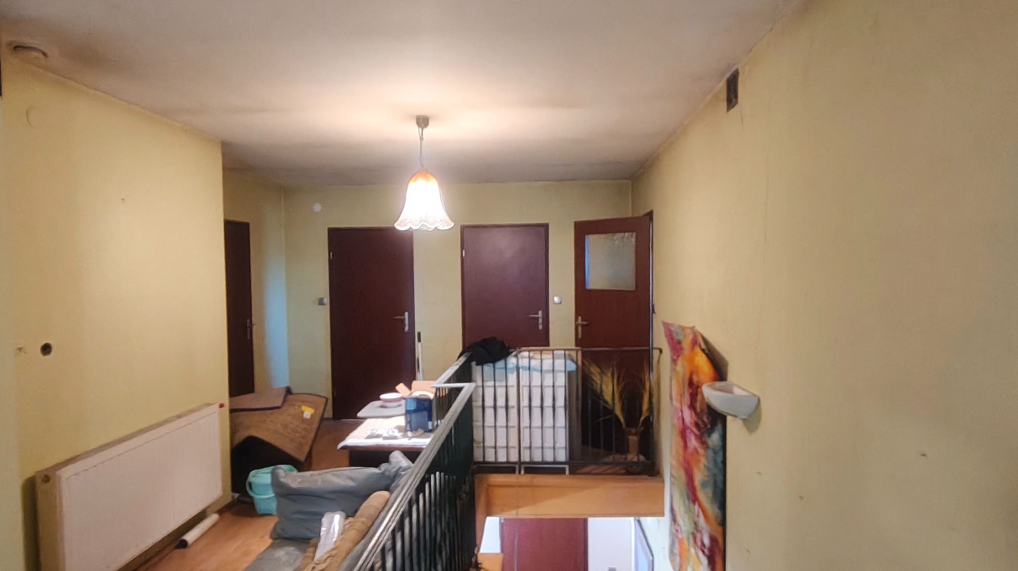 Korytarz w domu przed remontem: żółte ściany z widocznymi śladami użytkowania, trzy pary drzwi, lampa sufitowa, grzejnik i balustrada schodów. Widoczne niedoskonałości ścian.