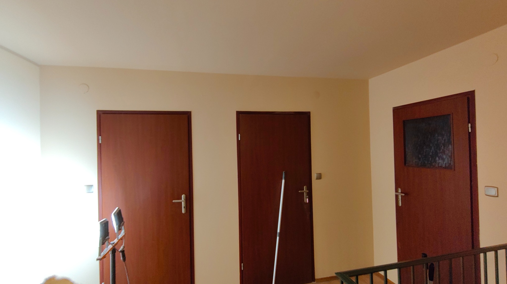 Korytarz po odświeżeniu ścian w odcieniach beżu, z trzema ciemnymi drzwiami. Widoczny sprzęt malarski i kij oparty o ścianę, sugerujący świeżo zakończone prace.