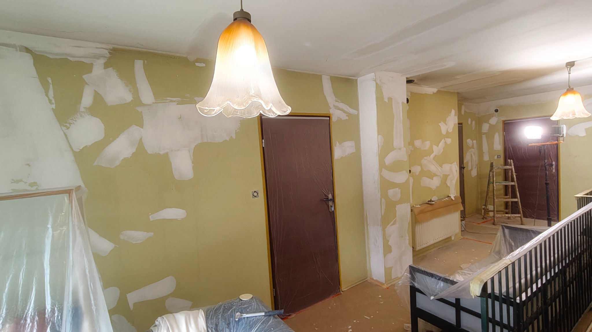 Pokój w trakcie szpachlowania: ściany z nałożonymi łatami gipsu, zabezpieczone drzwi folią, widoczna drabina i lampa wisząca. Prace wykończeniowe w trakcie.