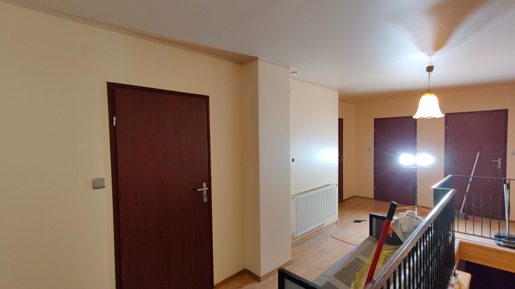 Korytarz po odświeżeniu: kremowe ściany, ciemne drzwi, widoczny grzejnik i lampa sufitowa. Świeżo pomalowane wnętrze z elementami wykończeniowymi.