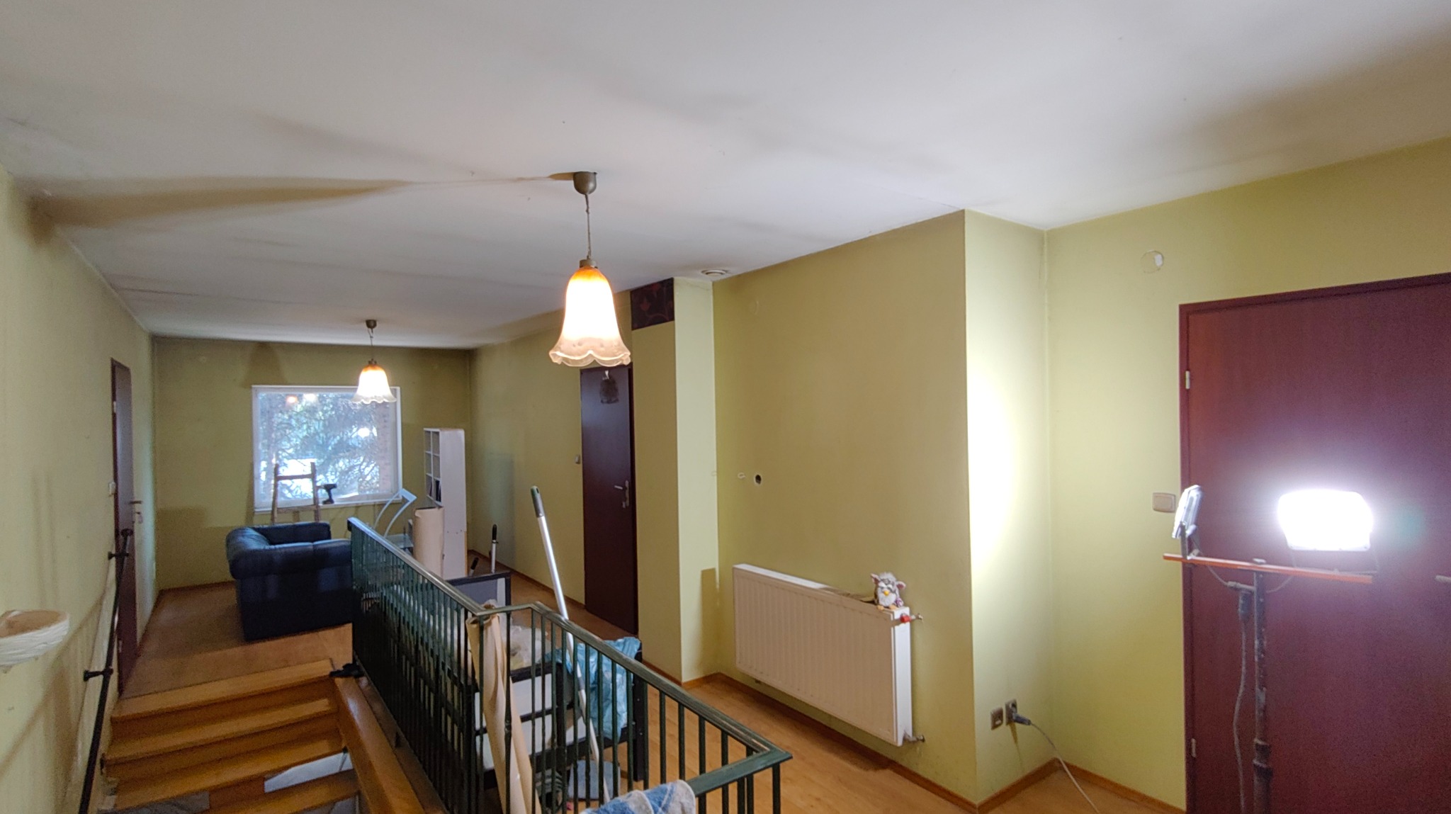 Wnętrze domu przed remontem: zielone ściany, plamy na suficie, widoczne schody i lampa wisząca. Na ścianie grzejnik i lampa robocza. Widok z góry na salon.