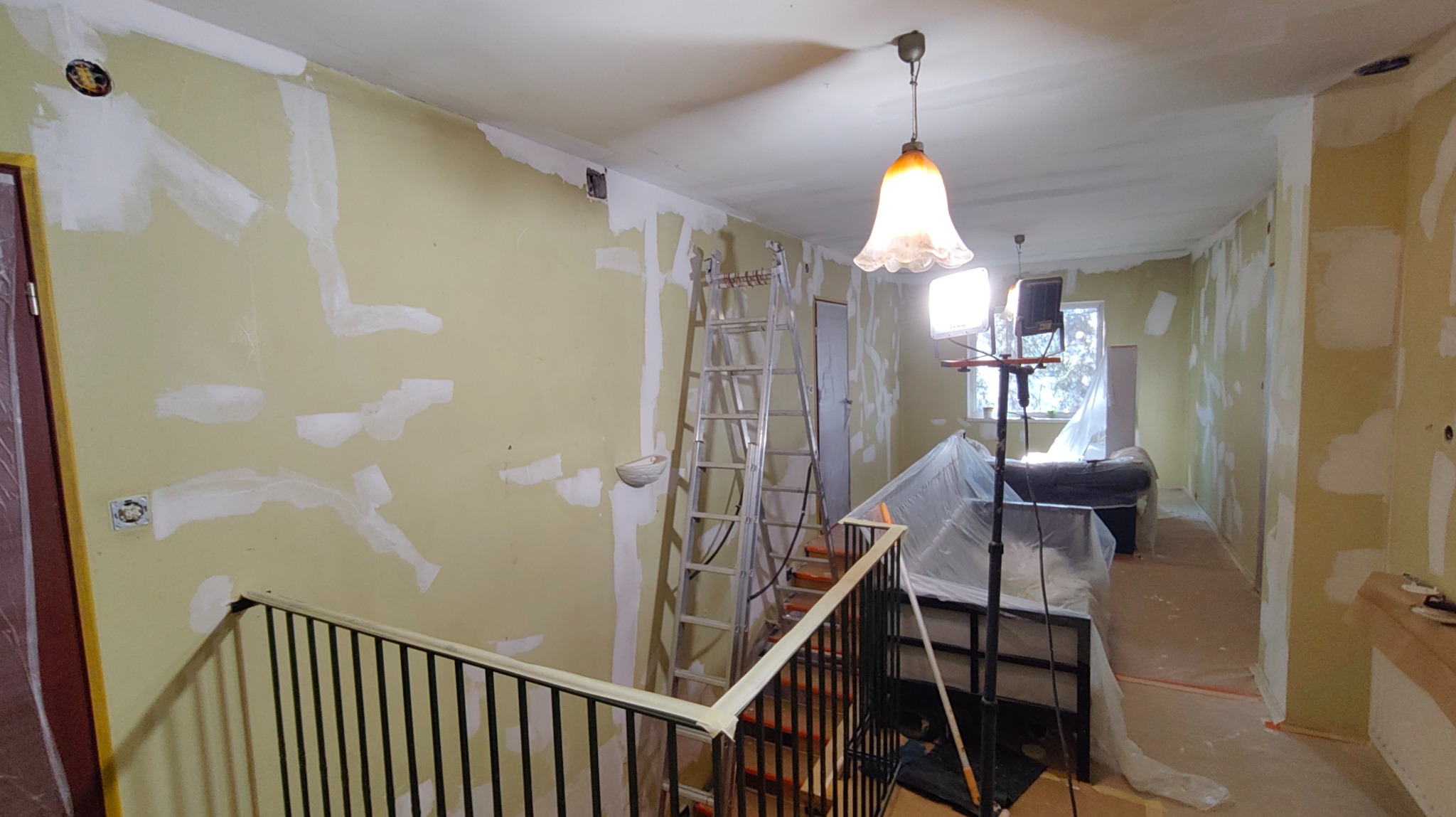 Wnętrze w trakcie szpachlowania: ściany z nałożoną gładzią, drabina, lampa oświetleniowa, zabezpieczone meble folią. Widok z góry schodów.