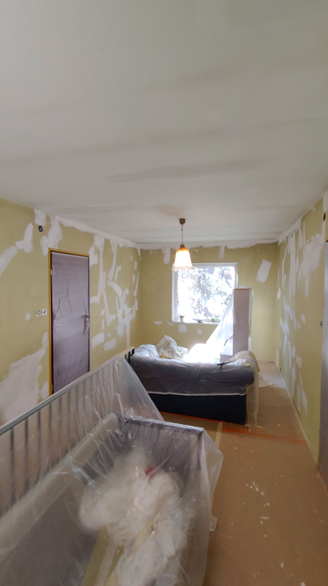 Pokój w trakcie prac przygotowawczych: ściany z nałożoną gładzią punktowo, meble zabezpieczone folią malarską, widoczne ślady po szpachlowaniu. Przygotowanie do malowania.