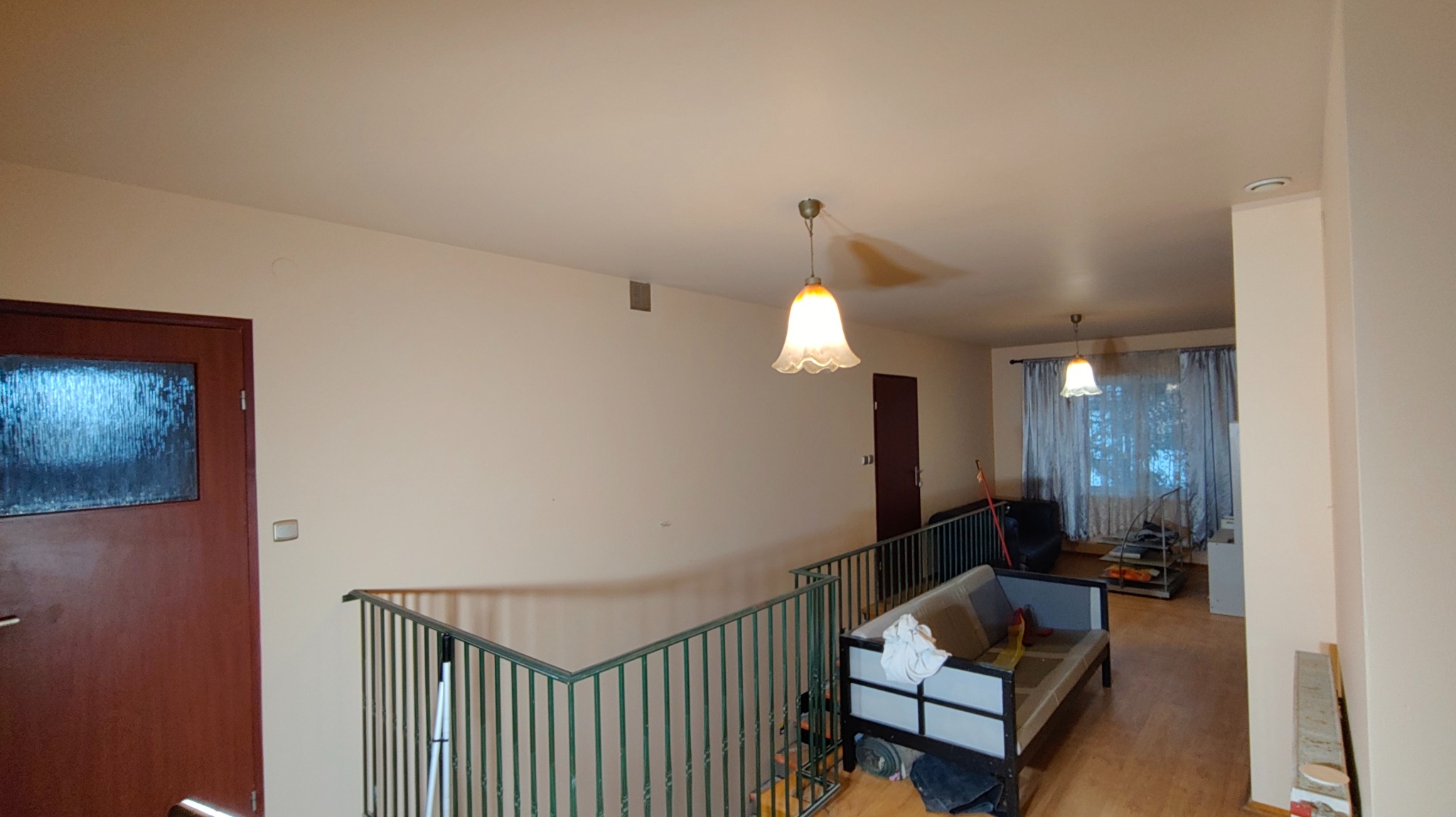 Jasne wnętrze domu przed odświeżeniem: beżowe ściany, drewniane drzwi i podłoga, widoczne plamy na suficie, metalowa barierka schodów. W tle okno z zasłonami.