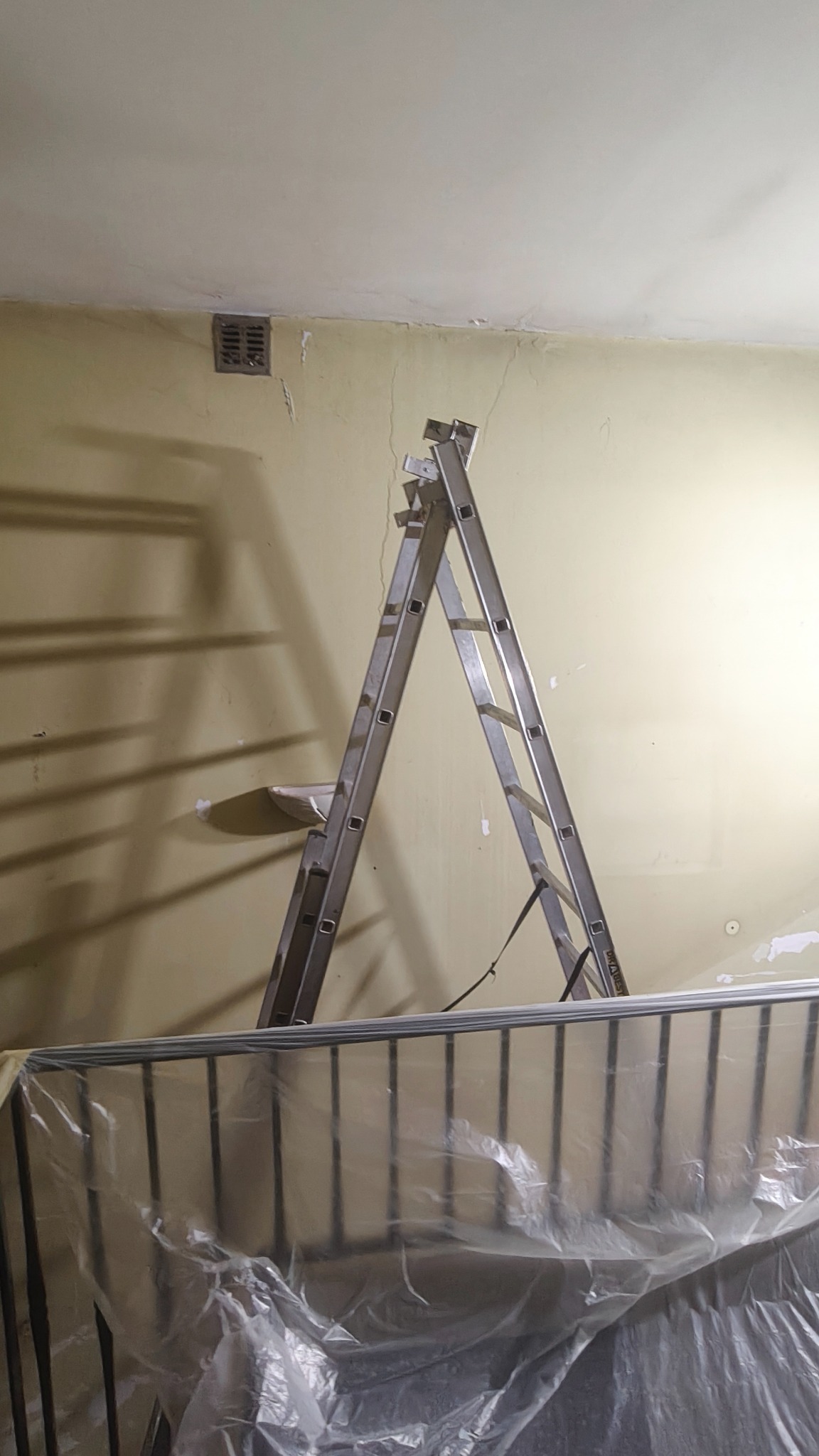 Przygotowania do remontu: drabina oparta o ścianę z widocznymi pęknięciami, zabezpieczone folią meble. Widoczny fragment kratki wentylacyjnej w górnej części ściany.