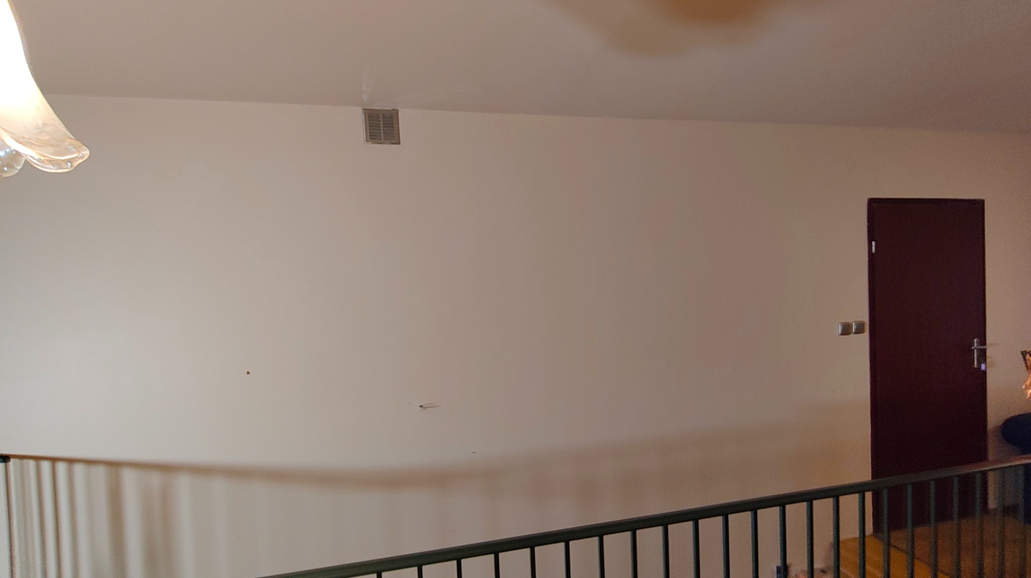 Jasna ściana po remoncie z widoczną kratką wentylacyjną pod sufitem i fragmentem balustrady na pierwszym planie. Ciemne drzwi po prawej stronie.