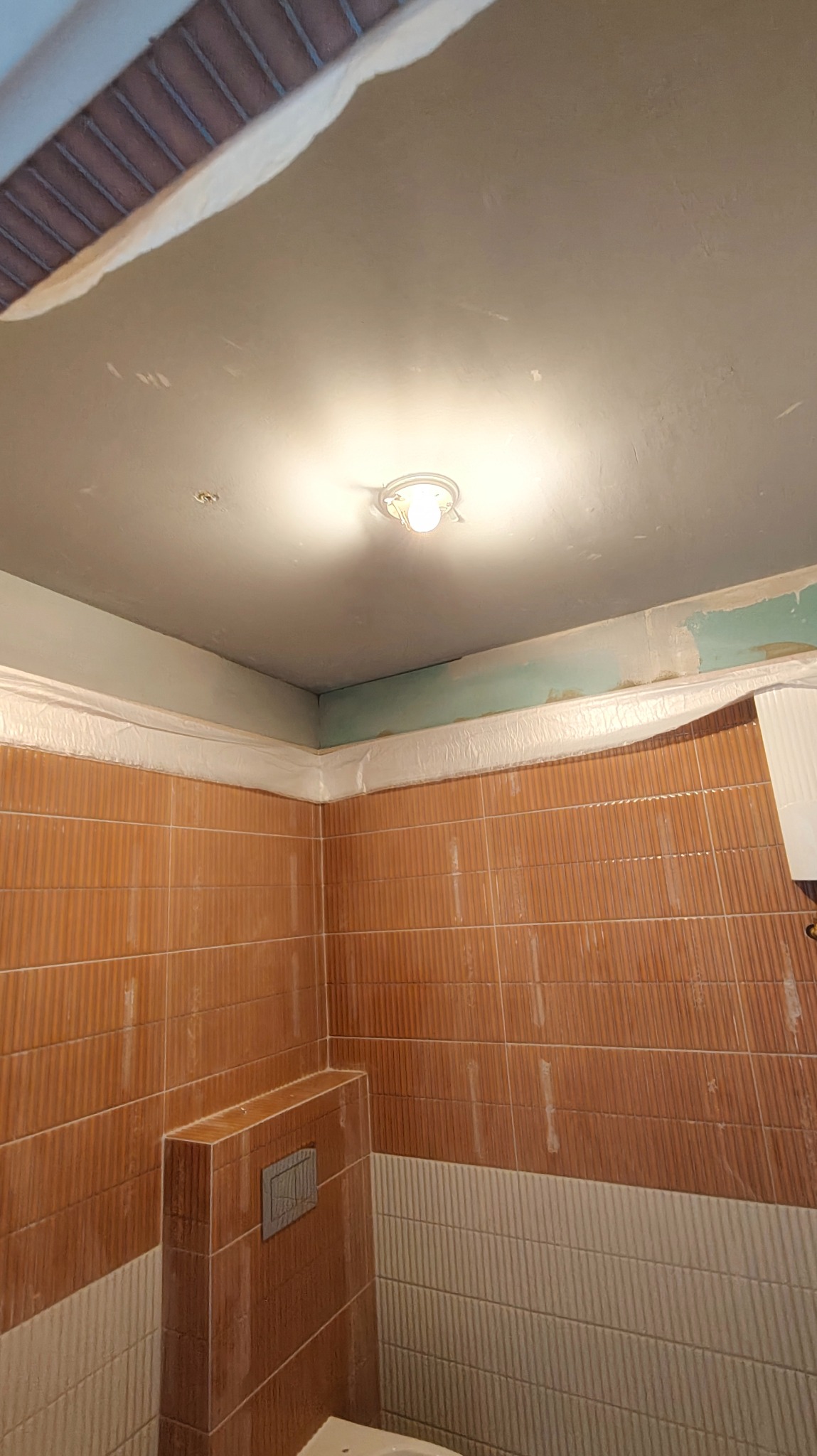 Łazienka w trakcie malowania sufitu na szaro, zabezpieczone folią płytki. Widoczne niedomalowane fragmenty i oświetlenie sufitowe. Prace wykończeniowe w toku.