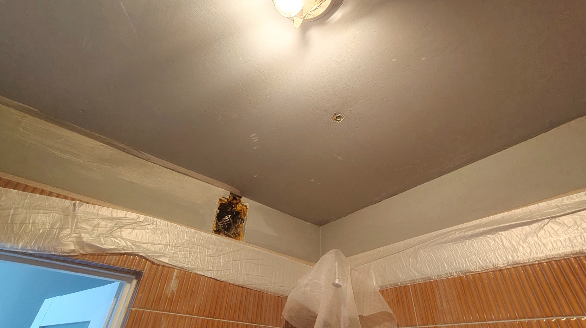 Szpachlowana sufitowa powierzchnia z widocznym otworem instalacyjnym, zabezpieczona folią ściana z brązowymi płytkami i oświetlenie sufitowe.