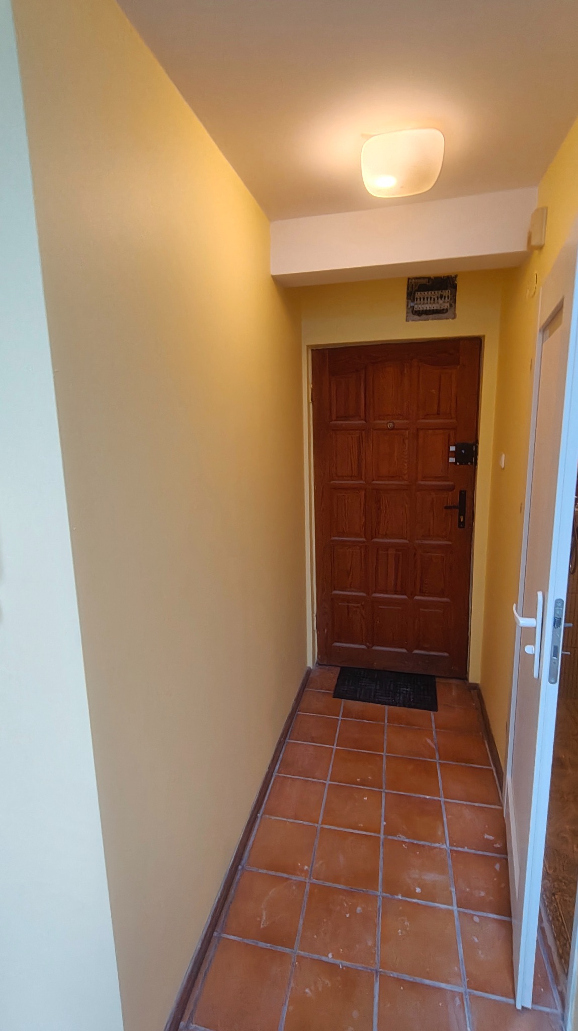 Korytarz po odświeżeniu: ściany w ciepłym odcieniu beżu, brązowe płytki na podłodze, drewniane drzwi wejściowe. Widoczne gładkie powierzchnie ścian i sufitu.