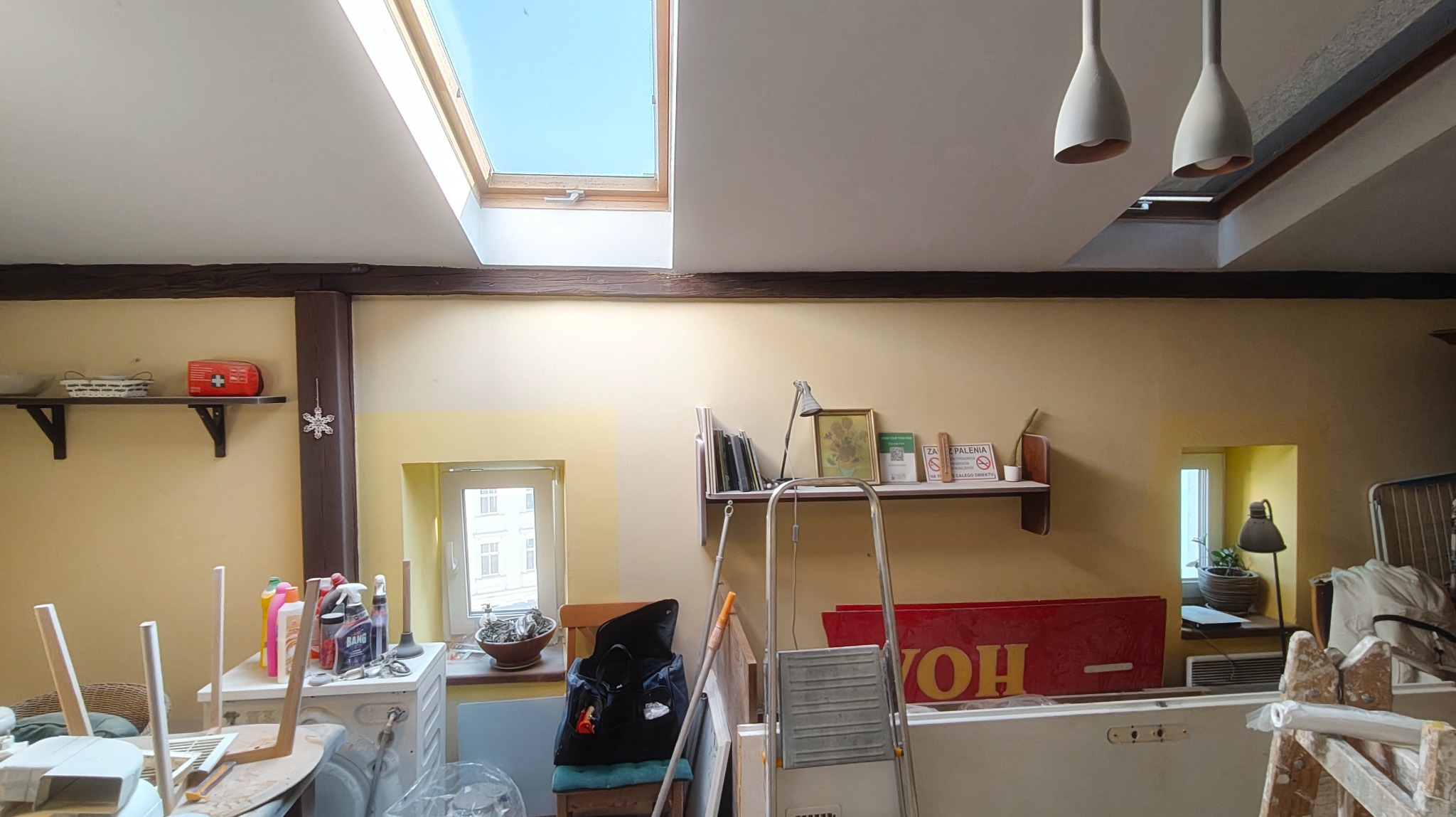 Wnętrze pokoju w trakcie odświeżania: ściany w odcieniach beżu, drabina malarska, okno dachowe i widoczne ślady prac remontowych. Częściowo pomalowana ściana.