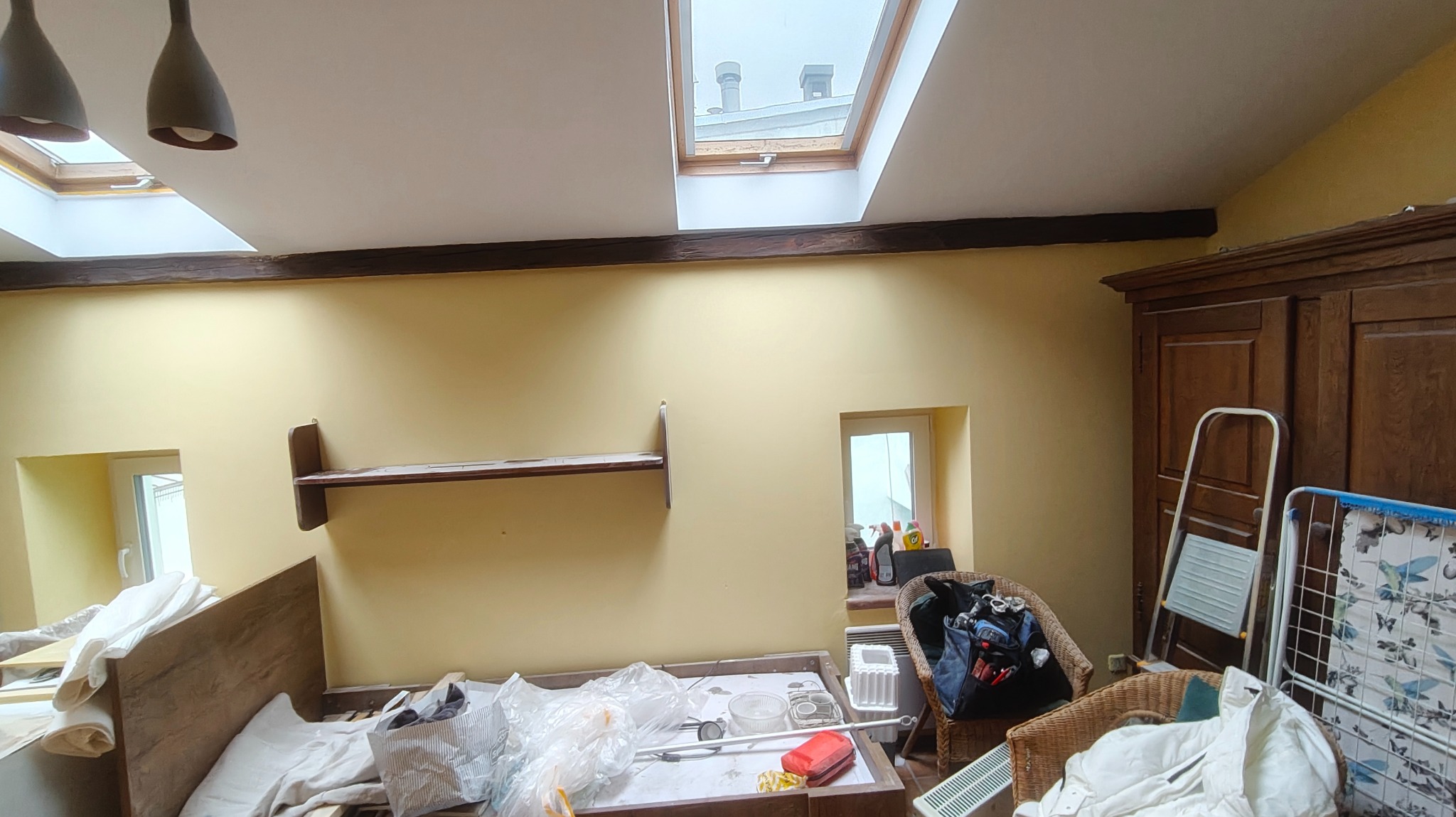 Pokój przed remontem: żółte ściany, drewniane belki stropowe, okno dachowe, meble i akcesoria przygotowane do zabezpieczenia przed pracami remontowymi.