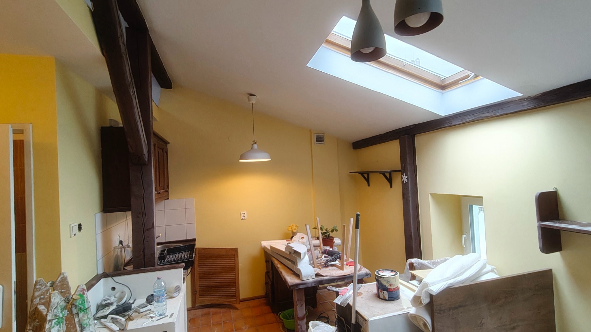 Żółte wnętrze w trakcie prac wykończeniowych: widoczne drewniane belki, okno dachowe, narzędzia i materiały malarskie. Przygotowanie do malowania ścian.