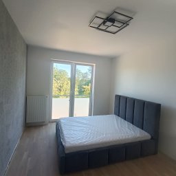 Artur Puchta - Nowoczesna sypialnia z szarą tapetą strukturalną, drewnianą podłogą i łóżkiem z szarym, tapicerowanym zagłówkiem, materac zabezpieczony folią, geometryczna lampa sufitowa.