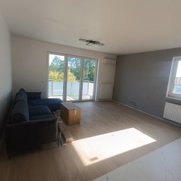 Artur Puchta - Wnętrze mieszkania w Łochowie po remoncie, z nową podłogą, szarą ścianą i rozpakowywaną sofą, widok na balkon.