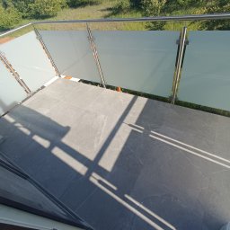 Artur Puchta - Wykończony balkon z szarą, kamienną podłogą i nowoczesną balustradą ze stali nierdzewnej i matowego szkła, widoczne cienie słoneczne na posadzce.
