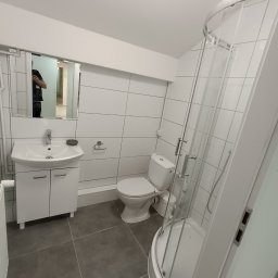 Artur Puchta - Wykończona łazienka z kabiną prysznicową, umywalką z szafką, toaletą i szarą podłogą z płytek, widoczna osoba odbijająca się w lustrze.