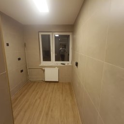 Artur Puchta - Wnętrze łazienki po remoncie z beżowymi płytkami, drewnopodobną podłogą, białym grzejnikiem i oknem z odbiciem osoby robiącej zdjęcie.