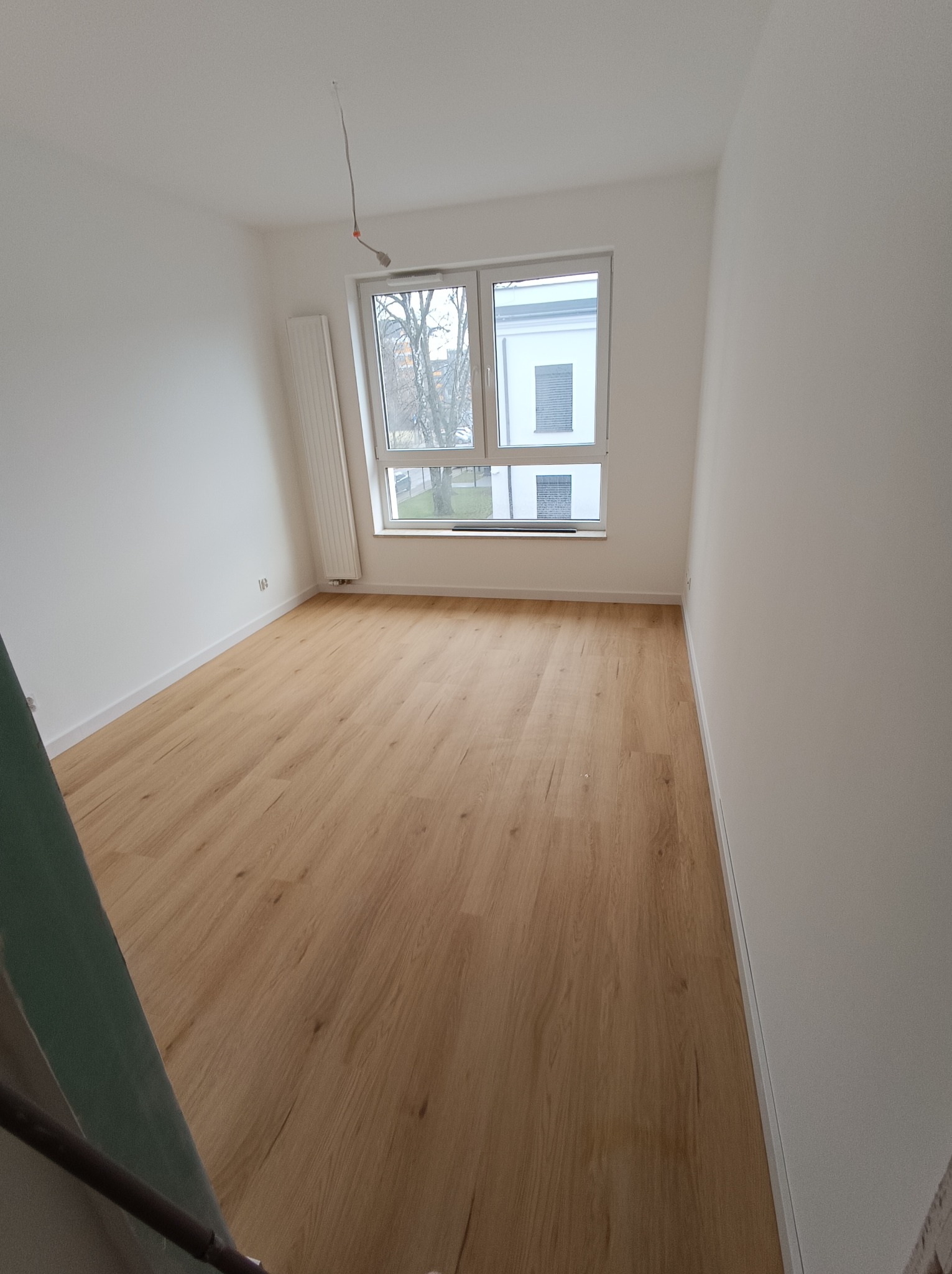 Pusty pokój po remoncie z drewnianą podłogą, białymi ścianami, grzejnikiem panelowym i oknem z widokiem na drzewa i budynek.