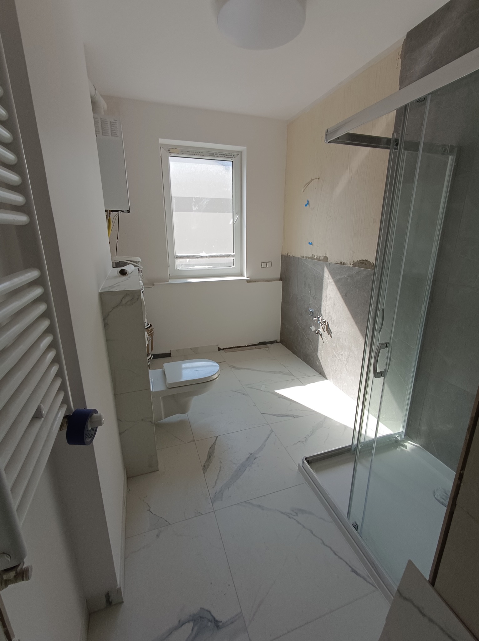 Wnętrze łazienki w trakcie remontu: widoczna kabina prysznicowa ze szklanymi drzwiami, biała toaleta, grzejnik drabinkowy, częściowo wykończone ściany z szarymi płytkami i białe marmurowe płytki...