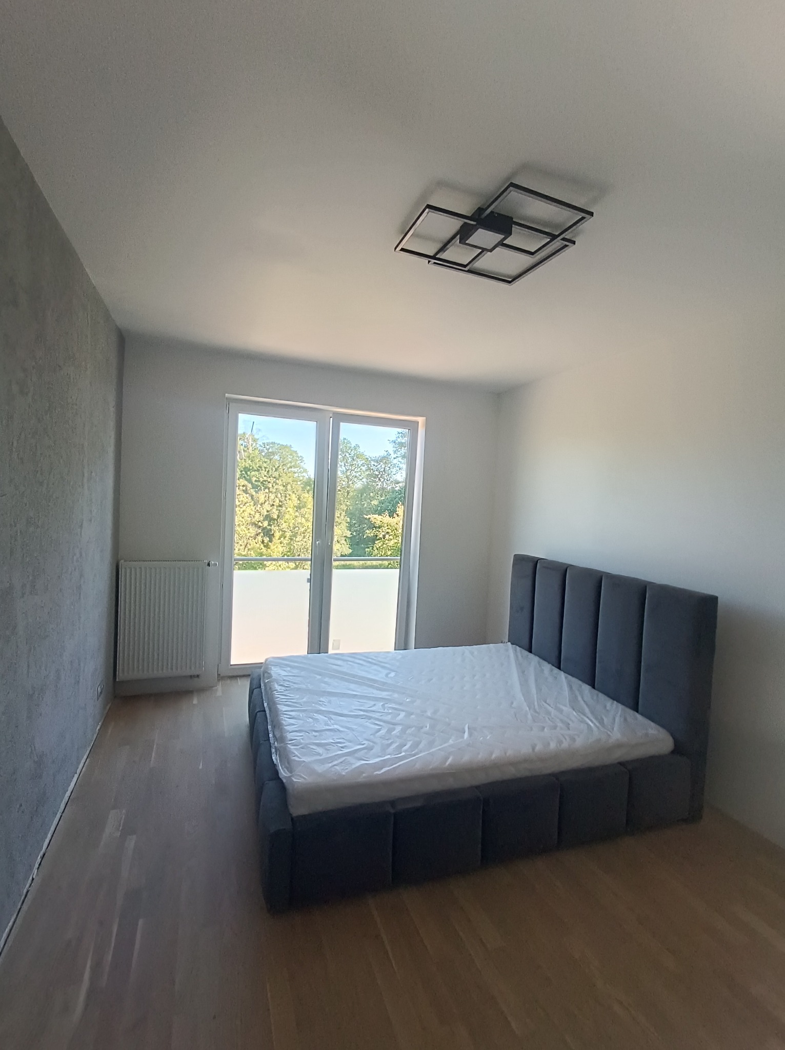 Nowoczesna sypialnia z szarą tapetą strukturalną, drewnianą podłogą i łóżkiem z szarym, tapicerowanym zagłówkiem, materac zabezpieczony folią, geometryczna lampa sufitowa.