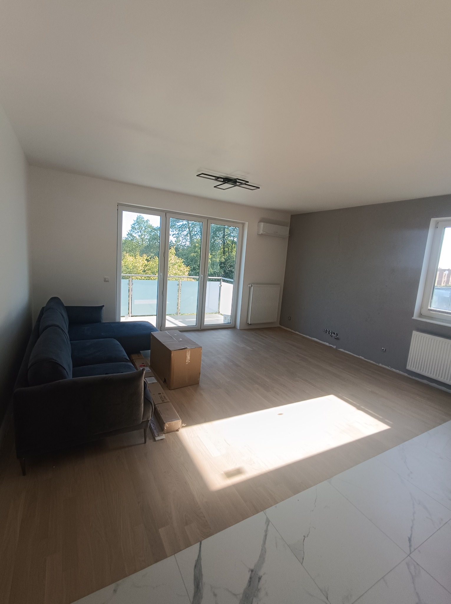 Wnętrze mieszkania w Łochowie po remoncie, z nową podłogą, szarą ścianą i rozpakowywaną sofą, widok na balkon.