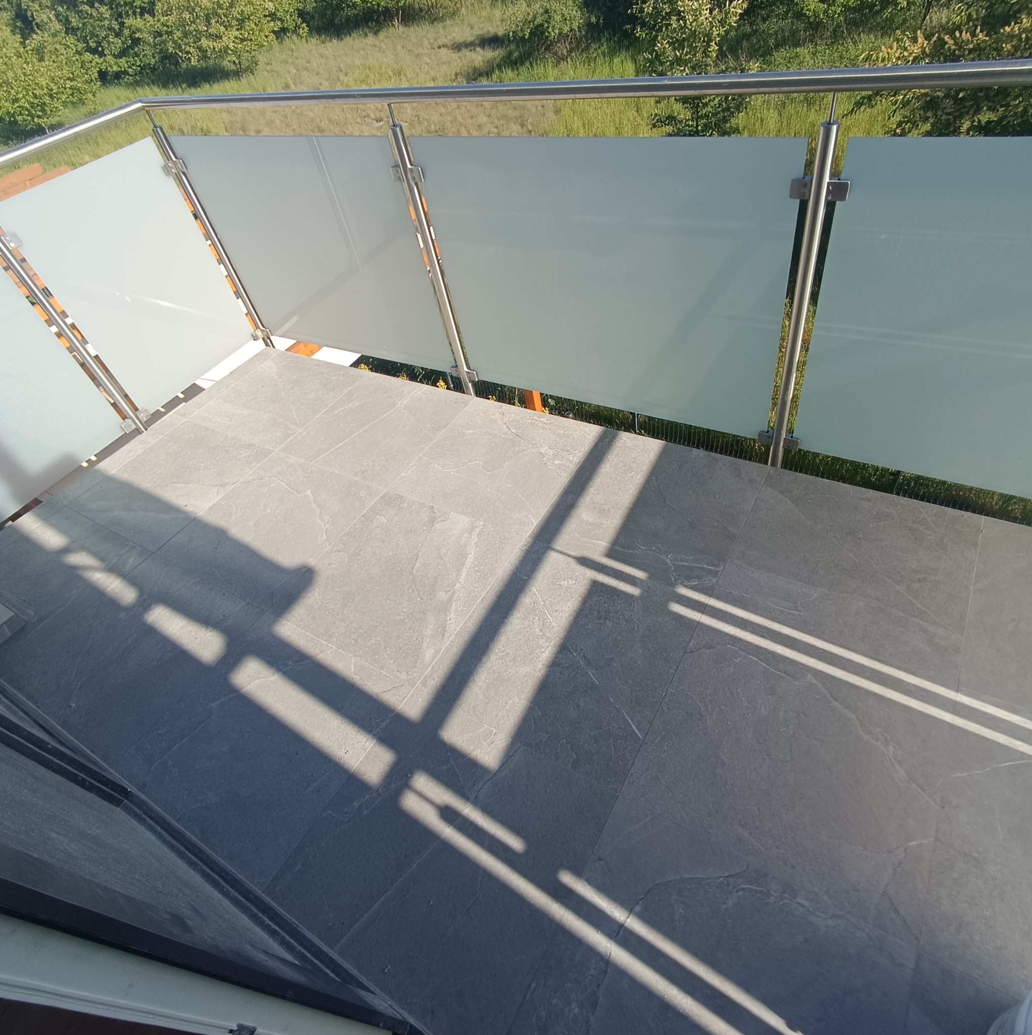 Wykończony balkon z szarą, kamienną podłogą i nowoczesną balustradą ze stali nierdzewnej i matowego szkła, widoczne cienie słoneczne na posadzce.
