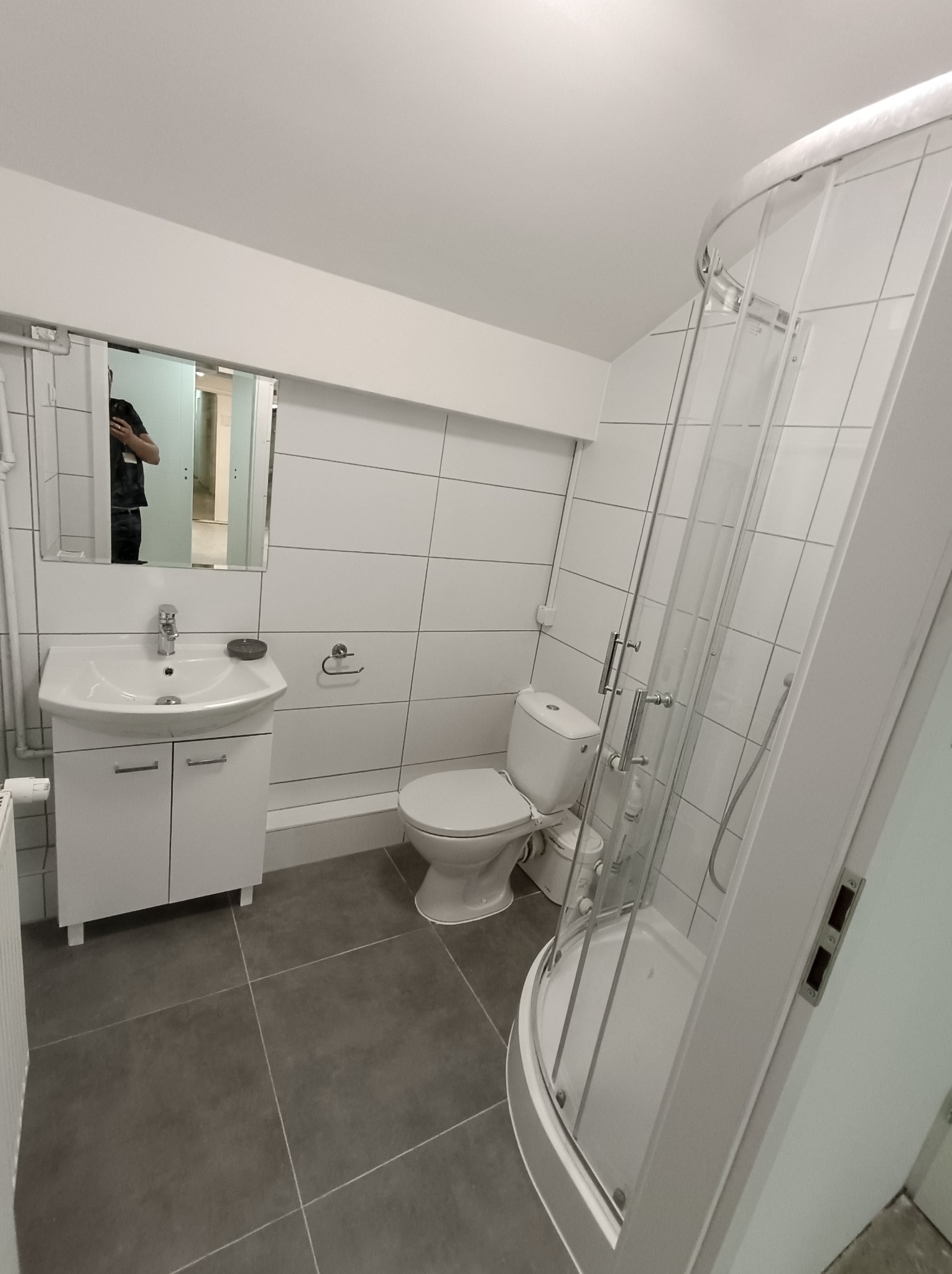 Wykończona łazienka z kabiną prysznicową, umywalką z szafką, toaletą i szarą podłogą z płytek, widoczna osoba odbijająca się w lustrze.