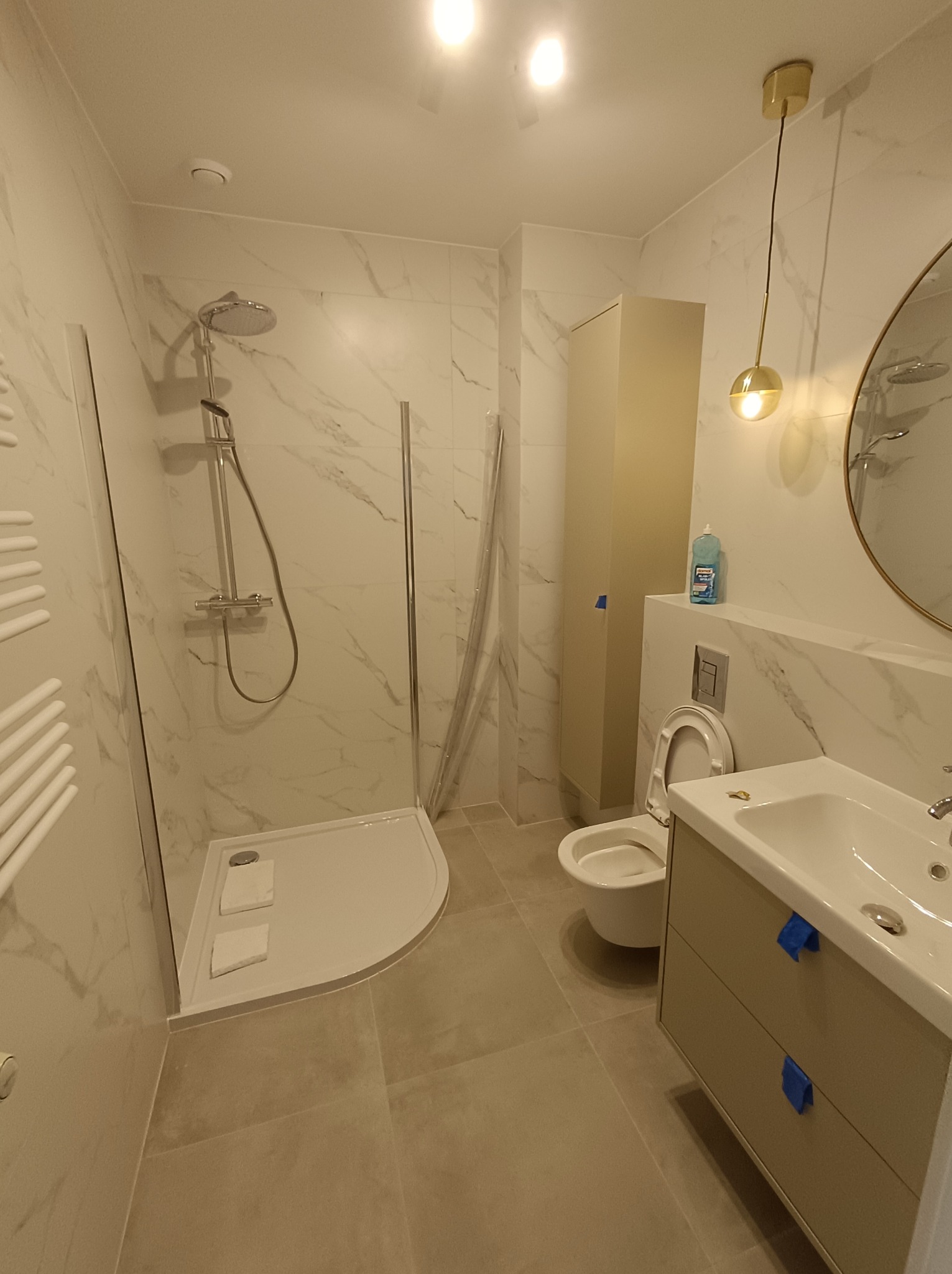 Nowoczesna łazienka z kabiną prysznicową typu walk-in, umywalką z szafką w beżowym odcieniu i toaletą; marmurowe płytki na ścianach, szklana ścianka prysznicowa, złota lampa wisząca i okrągłe lustro.