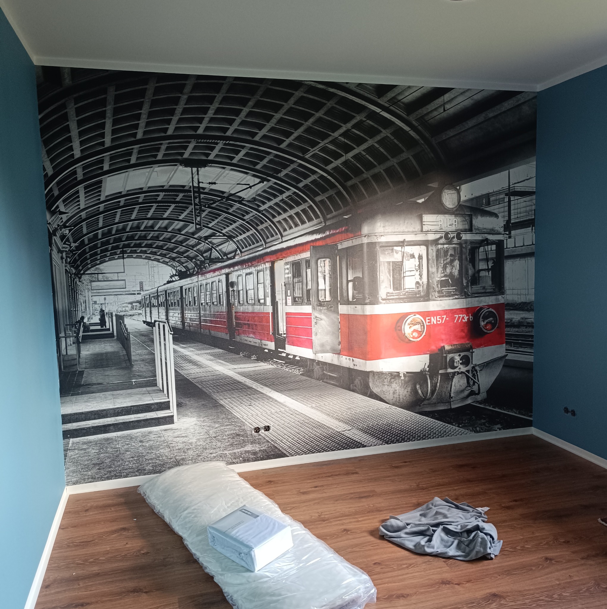 Fototapeta z motywem pociągu EN57 na ścianie w pokoju o niebieskich ścianach i drewnianej podłodze, na podłodze leżą zapakowane kołdry.