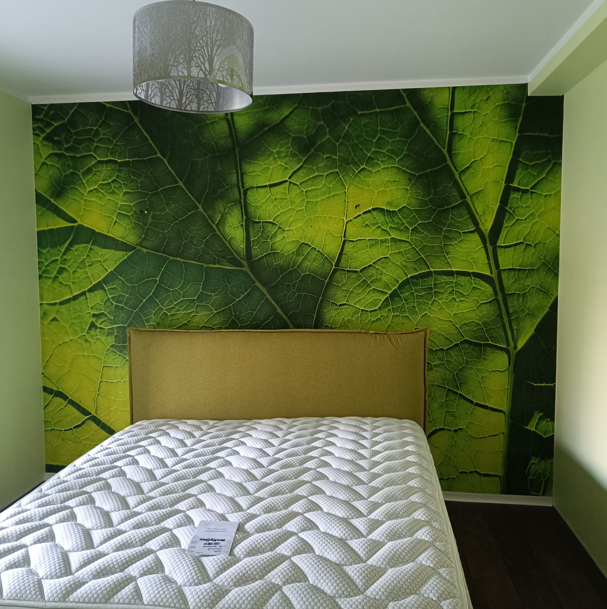 Sypialnia z łóżkiem z kremowym zagłówkiem i białym materacem, na ścianie fototapeta z motywem dużego zielonego liścia, nad łóżkiem lampa z abażurem z motywem drzew.