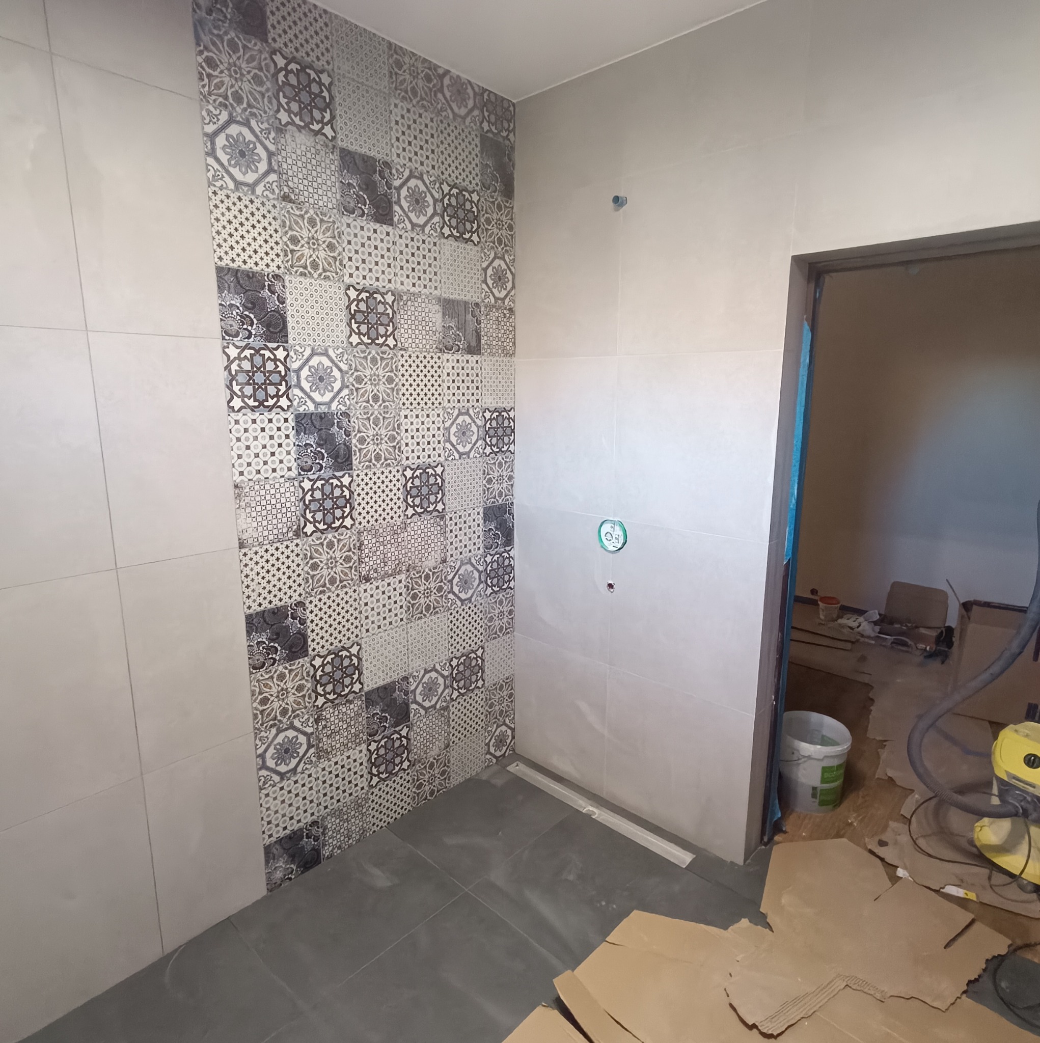 Fragment łazienki w trakcie prac wykończeniowych, widoczne płytki patchwork na ścianie, szare płytki podłogowe, odpływ liniowy w posadzce oraz narzędzia i materiały budowlane w tle.