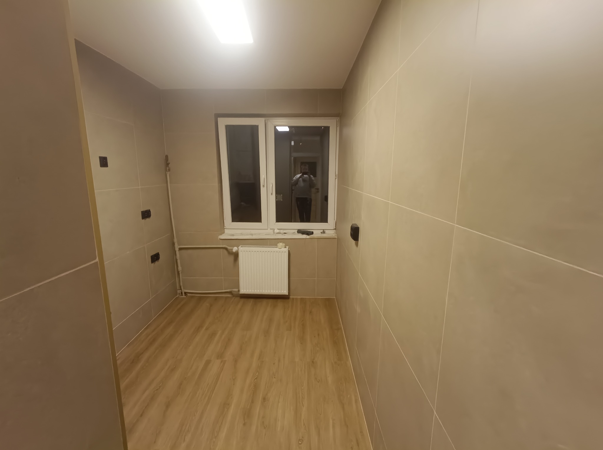 Wnętrze łazienki po remoncie z beżowymi płytkami, drewnopodobną podłogą, białym grzejnikiem i oknem z odbiciem osoby robiącej zdjęcie.