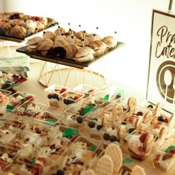 Praski Catering - Stół cateringowy z deserami w małych pojemnikach, kanapkami na paterach i logo firmy 'Praski Catering'. Jasne oświetlenie, ciepłe barwy.