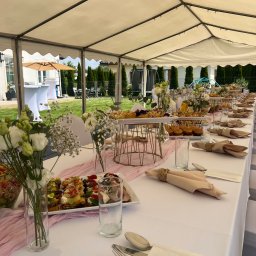 Praski Catering - Długi stół pod namiotem, nakryty białym obrusem, z dekoracjami kwiatowymi, zastawą i przekąskami. Widok na ogród z zielenią i budynkiem w tle.