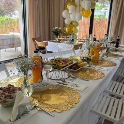 Praski Catering - Uroczysty stół z białym obrusem, złotymi podkładkami, sałatkami i napojami, udekorowany balonami w odcieniach bieli, złota i zieleni, ustawiony w jasnym wnętrzu z widokiem na ogród.