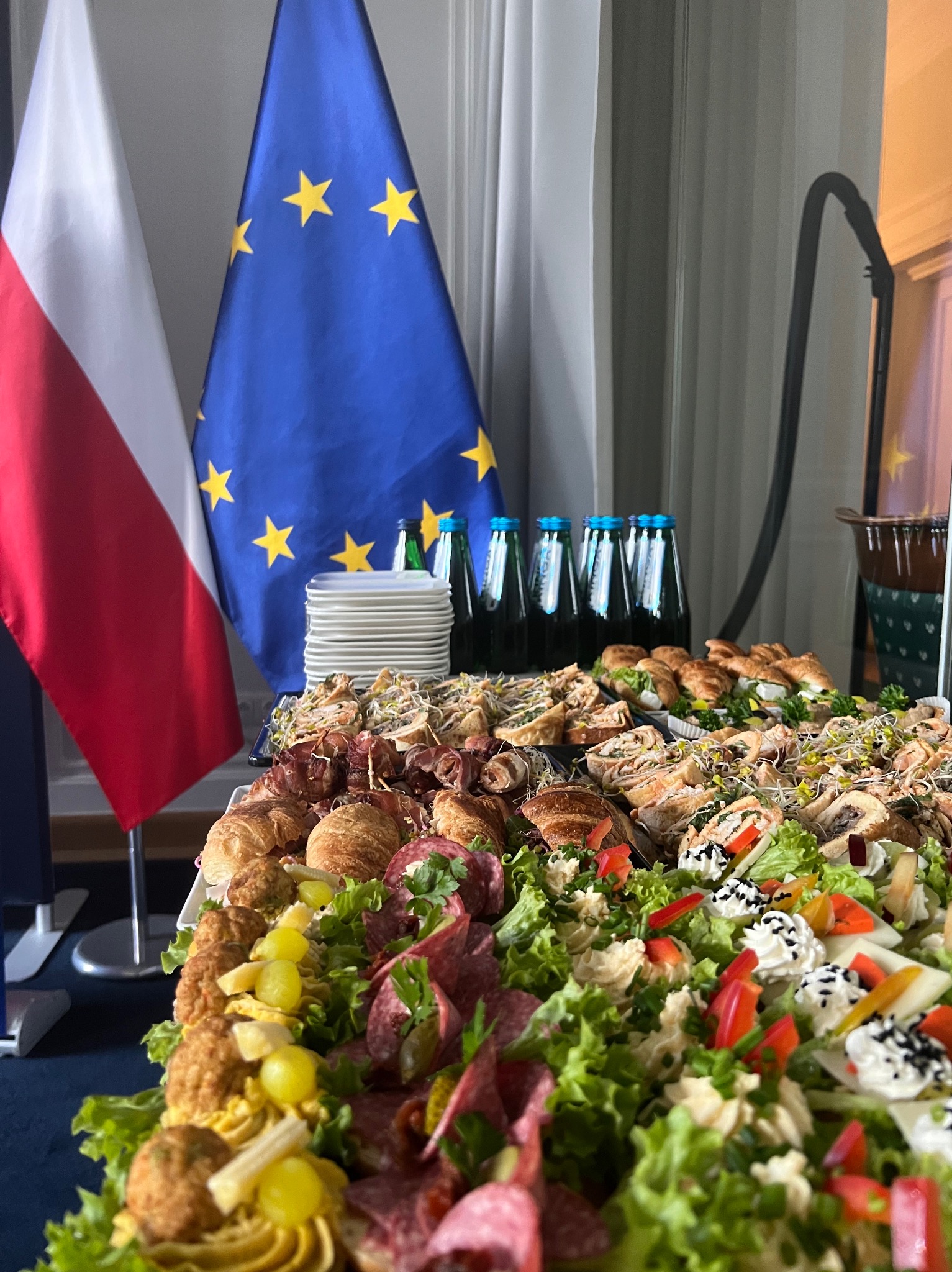 Stół cateringowy z przekąskami, na tle flagi Polski i Unii Europejskiej. Widoczne kanapki, sałatki i napoje. Uroczysty charakter wydarzenia.