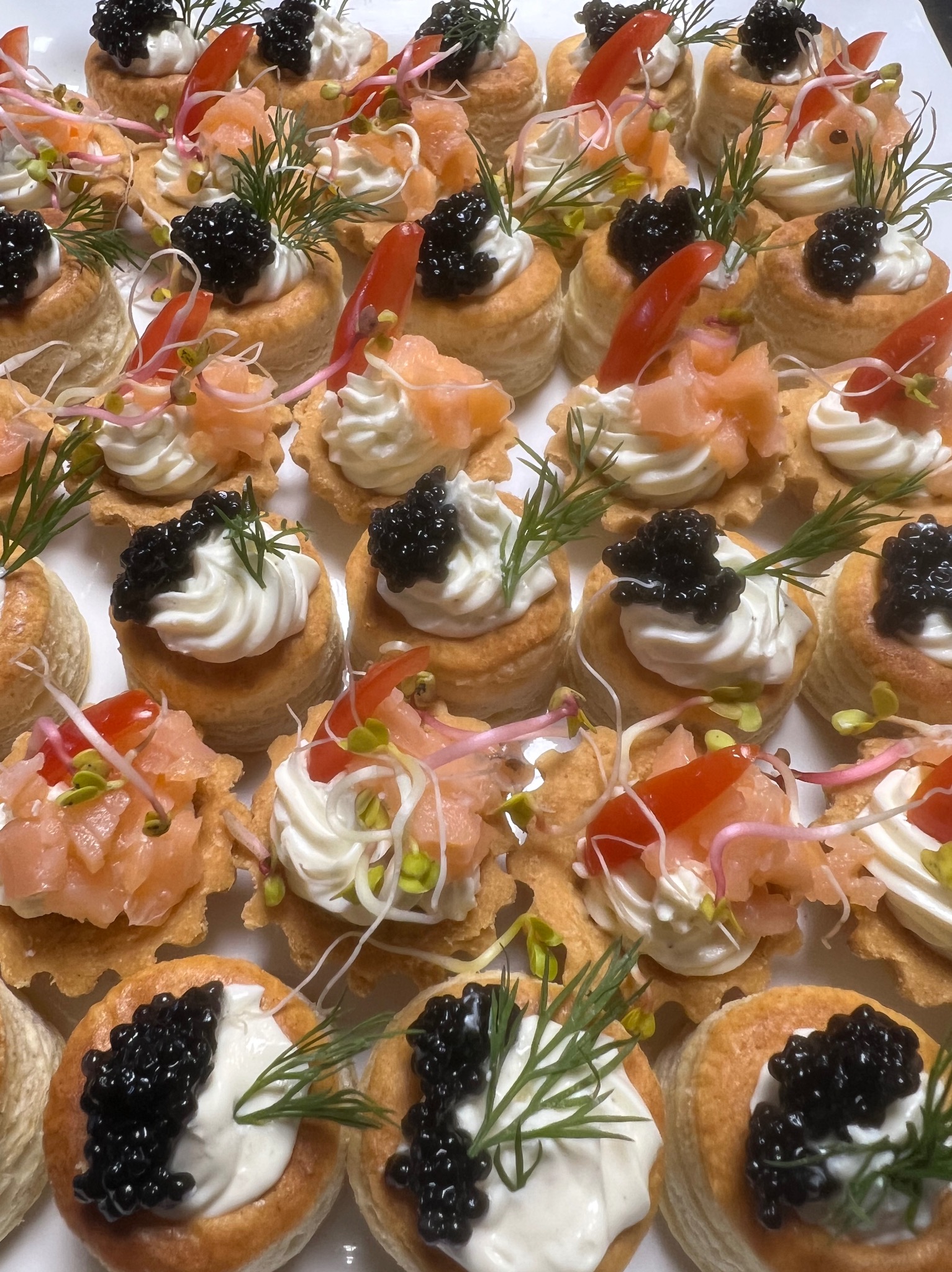 Eleganckie, różnorodne tartaletki: z kawiorem, łososiem i kremowym serkiem, udekorowane świeżymi ziołami i kiełkami. Idealne jako wykwintne przekąski.