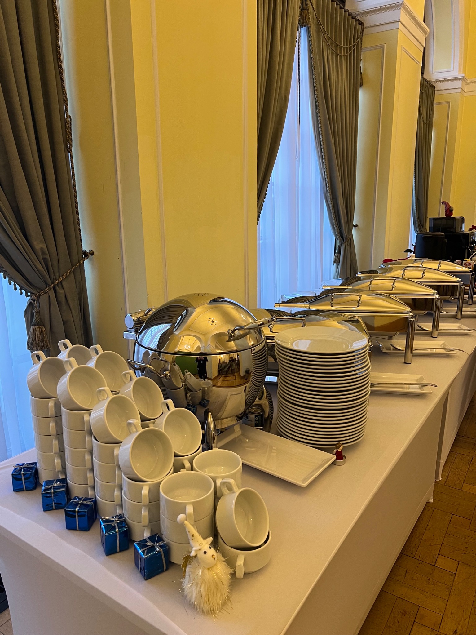 Elegancki bufet cateringowy: srebrne bemary, stosy białych naczyń, dekoracyjne prezenty i figurka, ustawione na stole z białym obrusem w wytwornym wnętrzu.